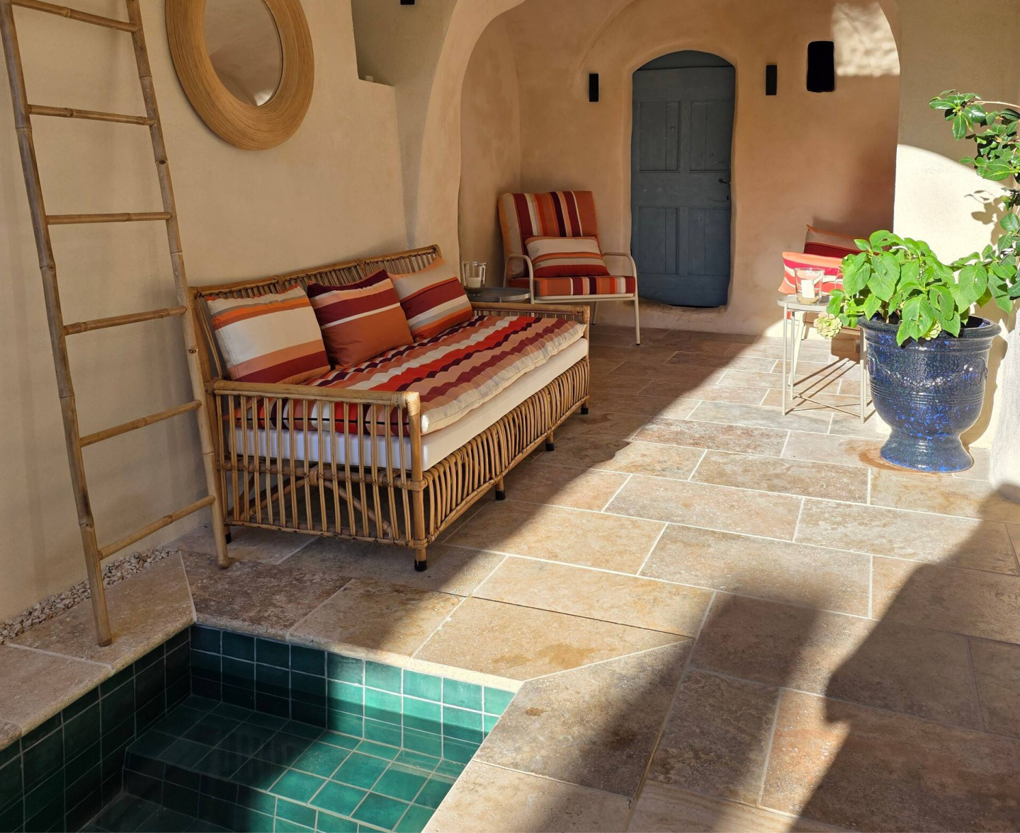 Sunlit cozy seating area with wicker sofa and chair featuring red and orange striped cushions, a small pool with green tiles, a large blue planter with green leafy plant, and a wooden ladder leaning against a beige wall.
