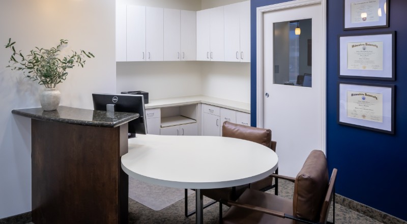 Small modern office with a white round table, two brown chairs, white cabinets, a computer, a vase with green branches, and framed diplomas on a blue wall.
