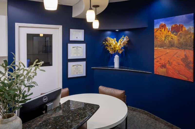Office room with dark blue walls, a round white table, leather chairs, a granite countertop, framed diplomas, a vase with yellow flowers, and landscape artwork of red rock formations.