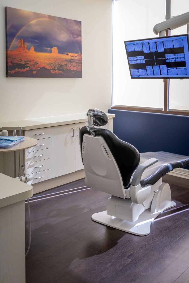 Modern dental examination room with a dental chair, X-ray images on a monitor, and a landscape painting on the wall.