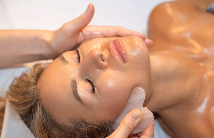 Woman receiving a facial massage with a gua sha tool, eyes closed and skin glowing.