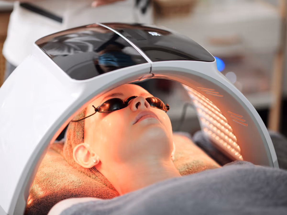 Person wearing protective goggles undergoing LED light therapy with a curved device emitting red light on their face.