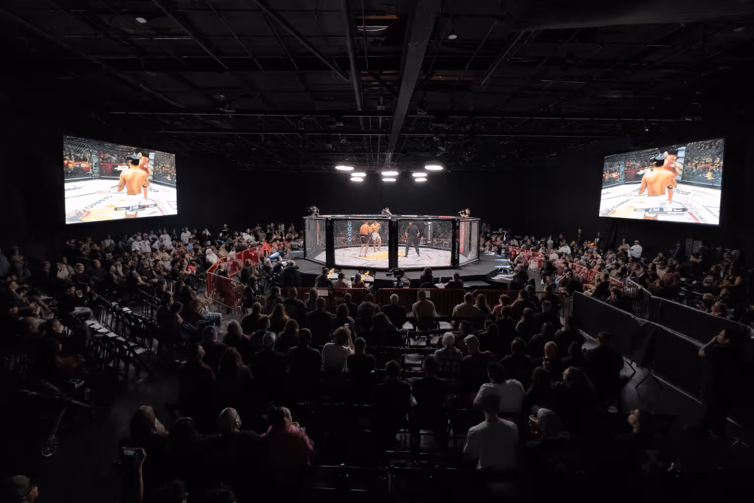 Packed indoor arena with audience watching a mixed martial arts fight inside a caged octagon, with large screens displaying the match at Thunder Studios