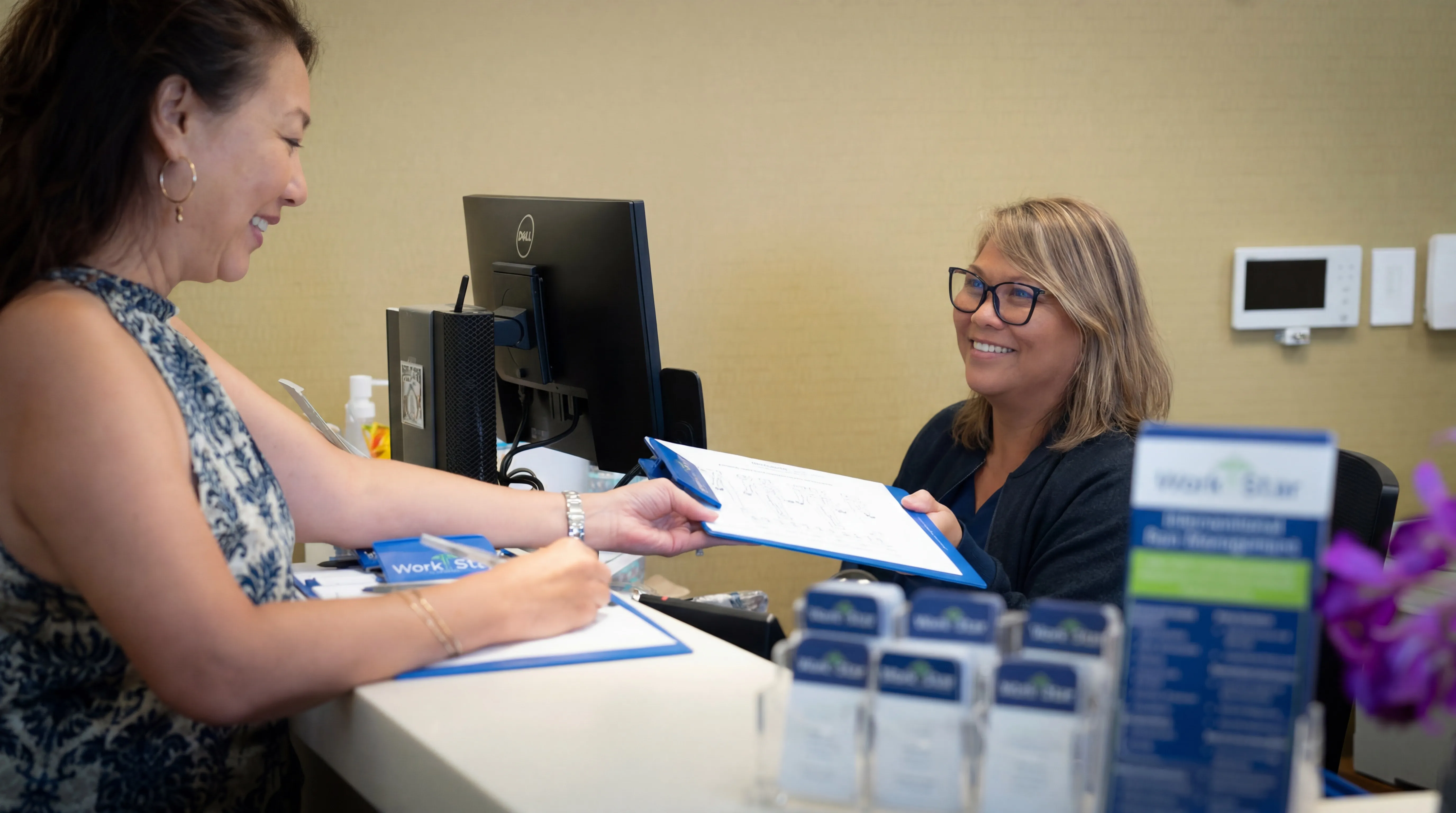 Woman smiles as she hands a clipboard with forms to a receptionist behind a desk in an office.