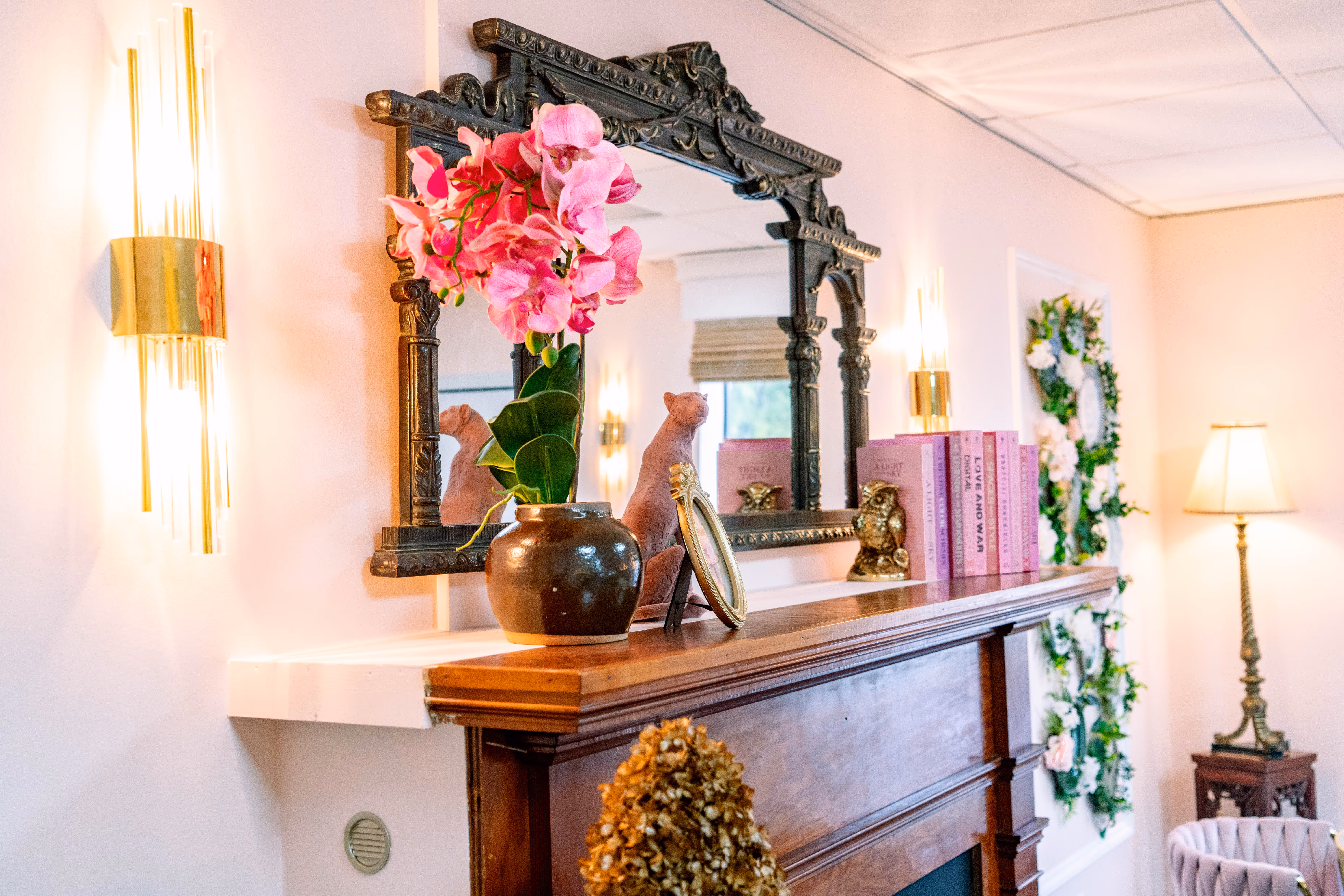 Mantelpiece with a dark ornate mirror, pink flowers in a vase, framed photo, pink books, and decorative owl, illuminated by wall sconces and a nearby lamp.