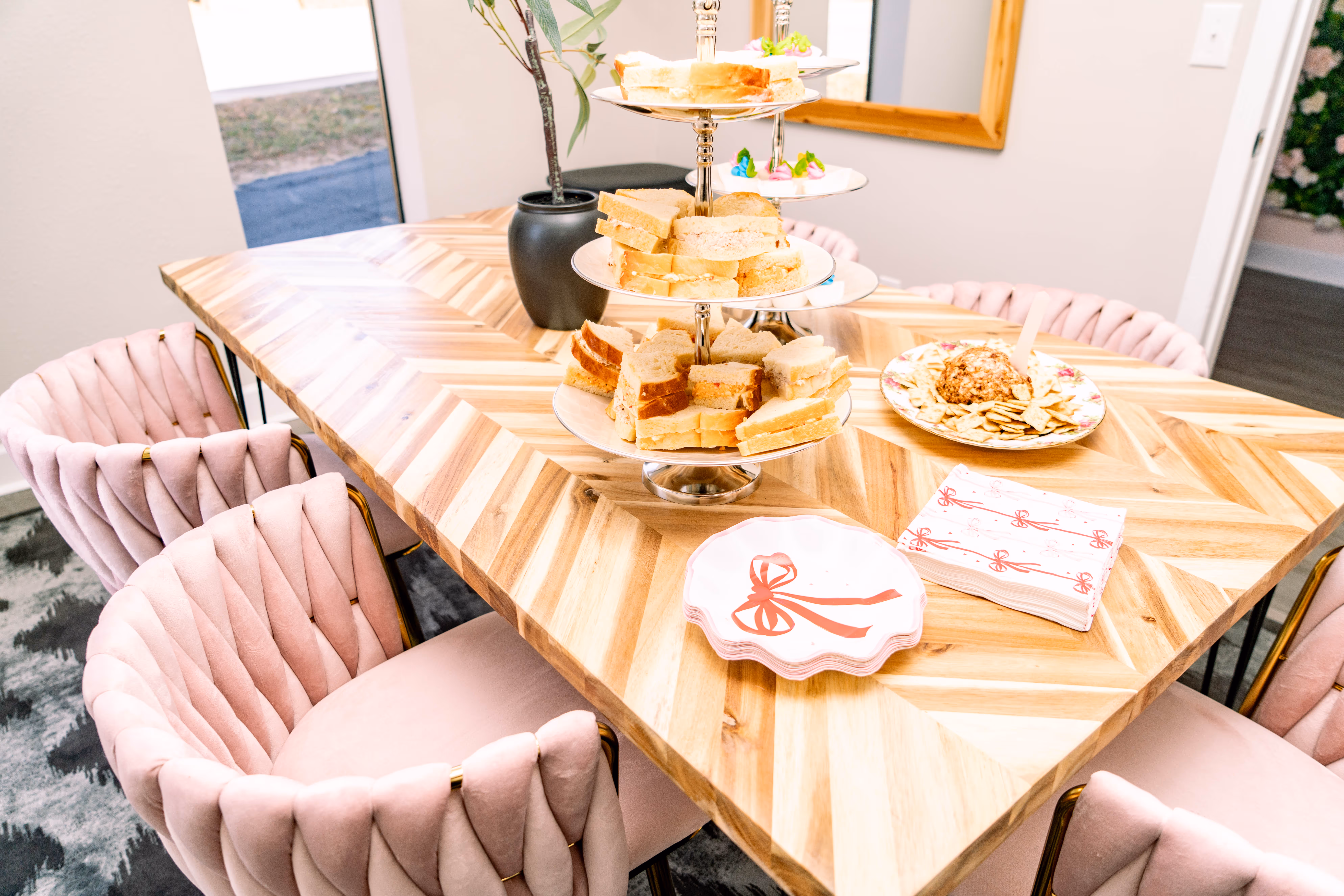 Wooden dining table with a three-tier tray of sandwiches, a plate of crackers and cheese ball, and pink velvet chairs around it.