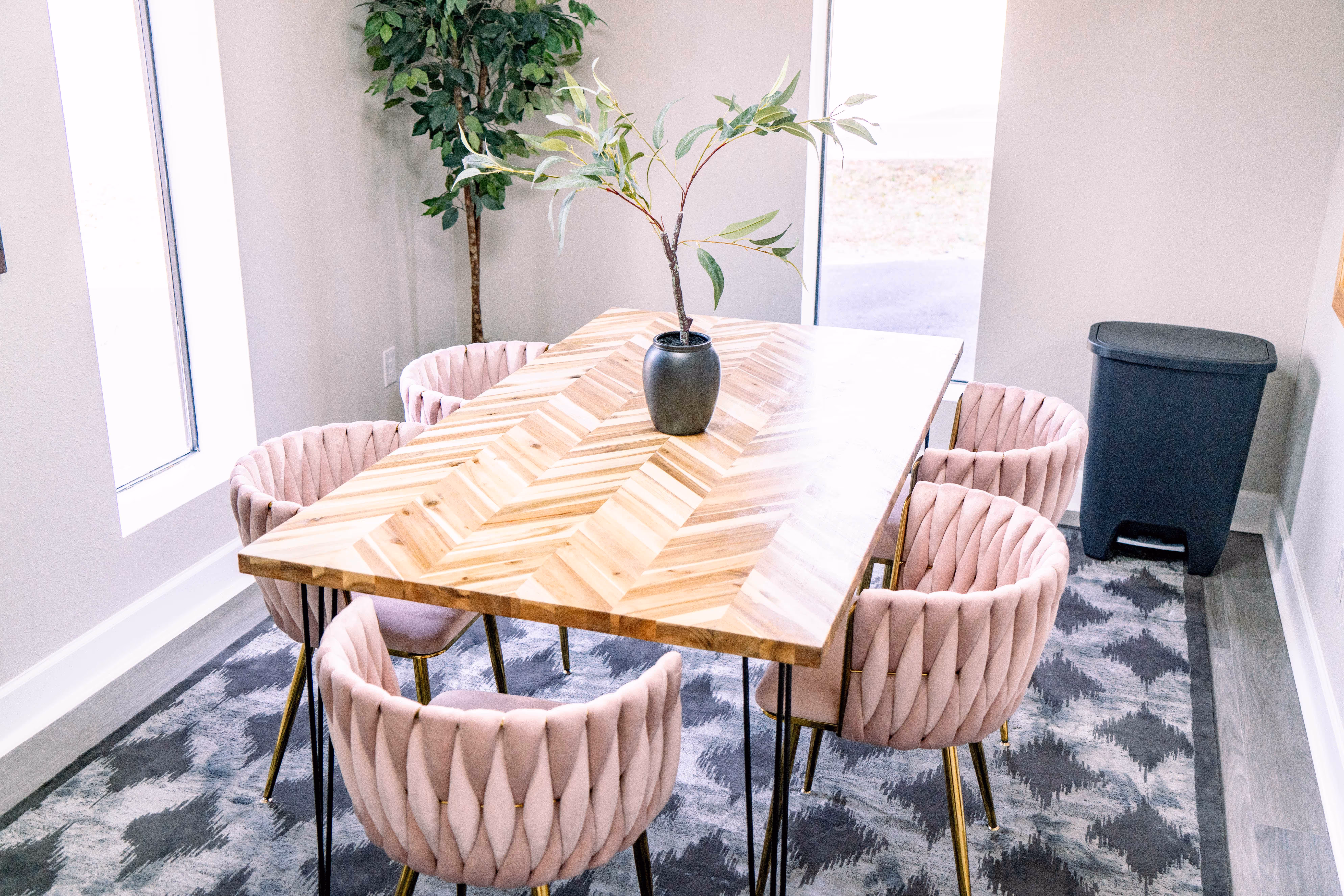 Dining room with a wooden chevron-patterned table, six pink velvet chairs with gold legs, a small plant in a black vase on the table, and a gray trash bin in the corner.