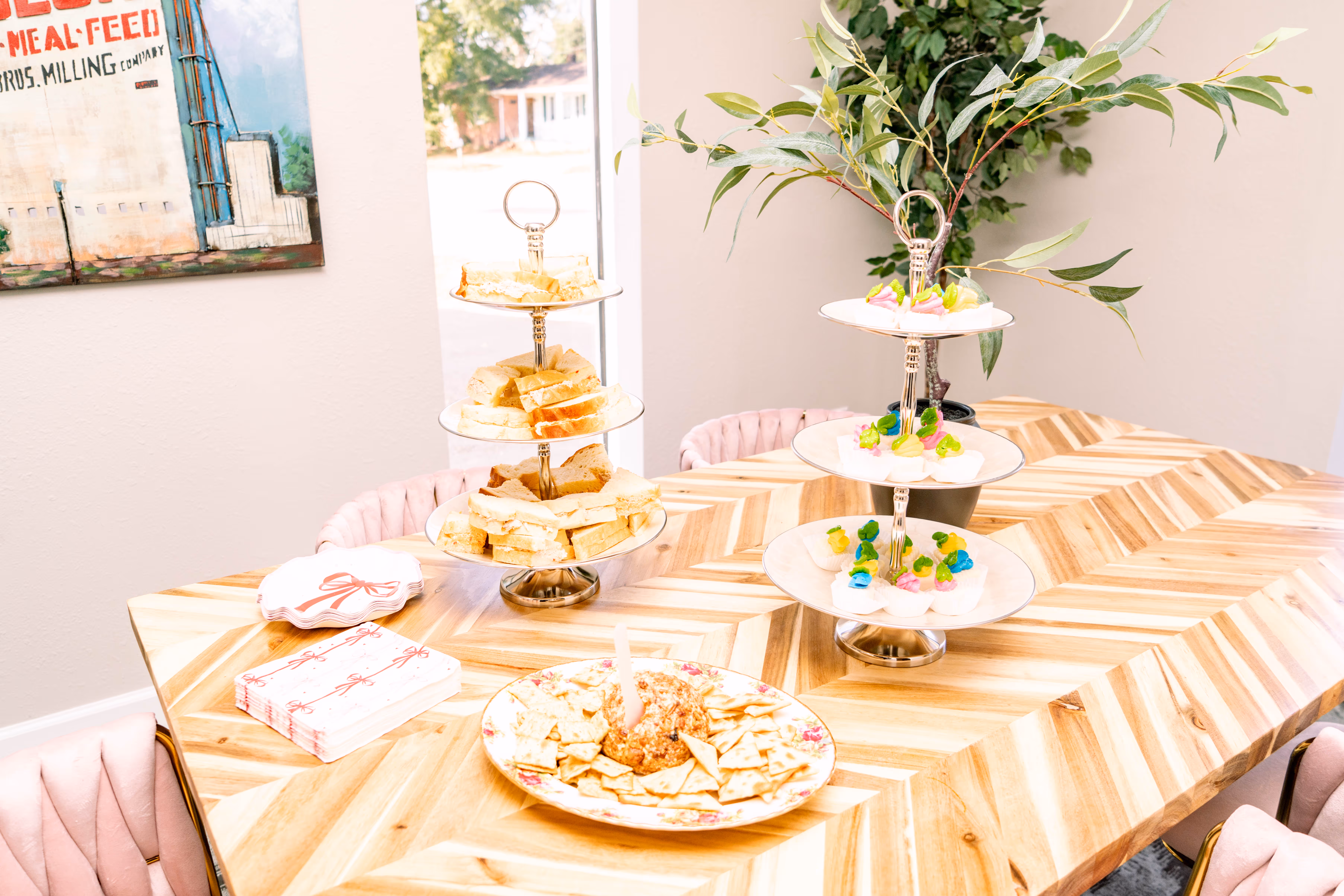 Wooden table with three-tiered trays holding sandwiches and decorated cupcakes, a plate of crackers with cheese spread, pink napkins, and pink chairs.