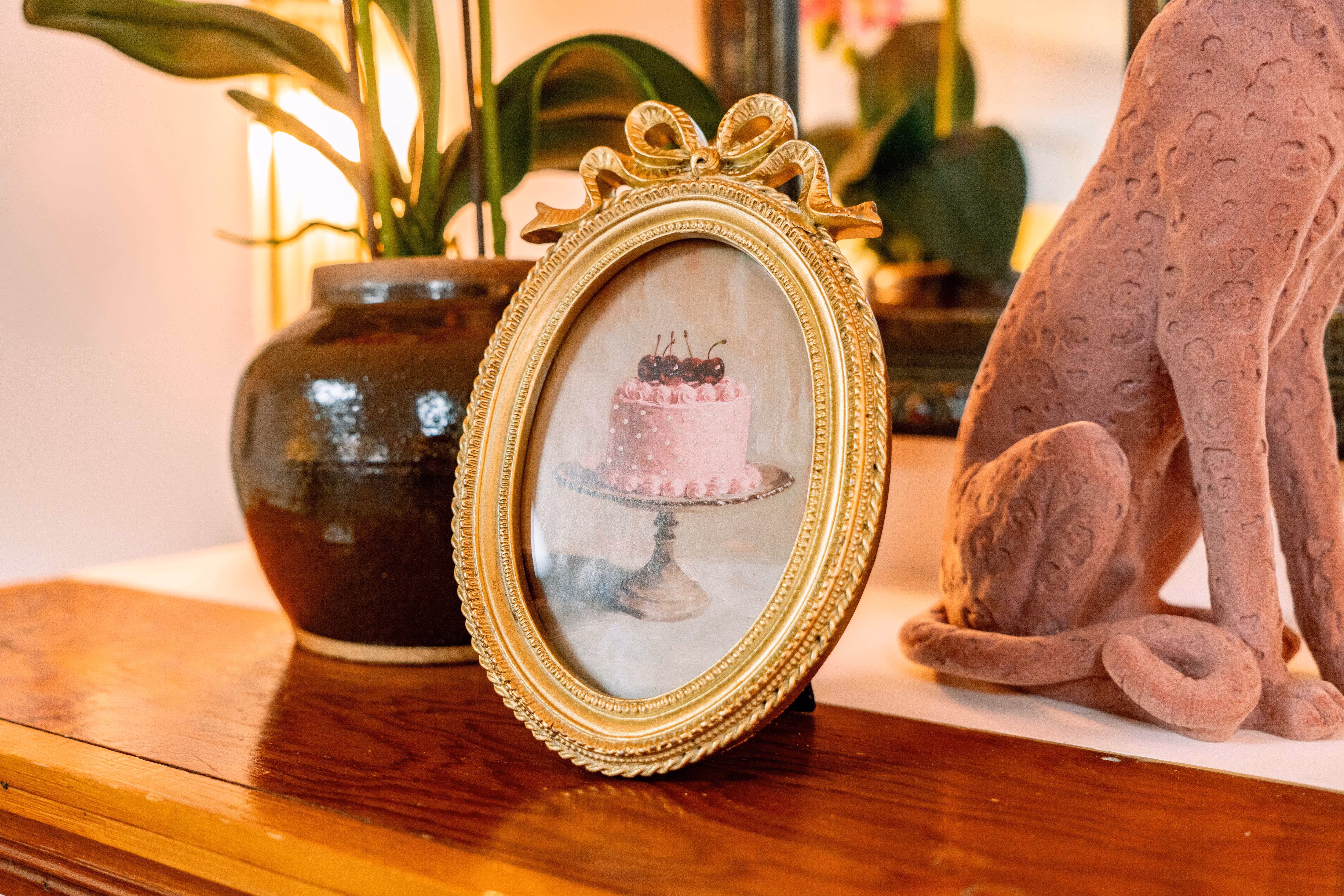 Ornate gold oval frame with a painting of a pink cake topped with cherries, placed on a wooden surface next to a brown ceramic pot and a pink leopard sculpture.