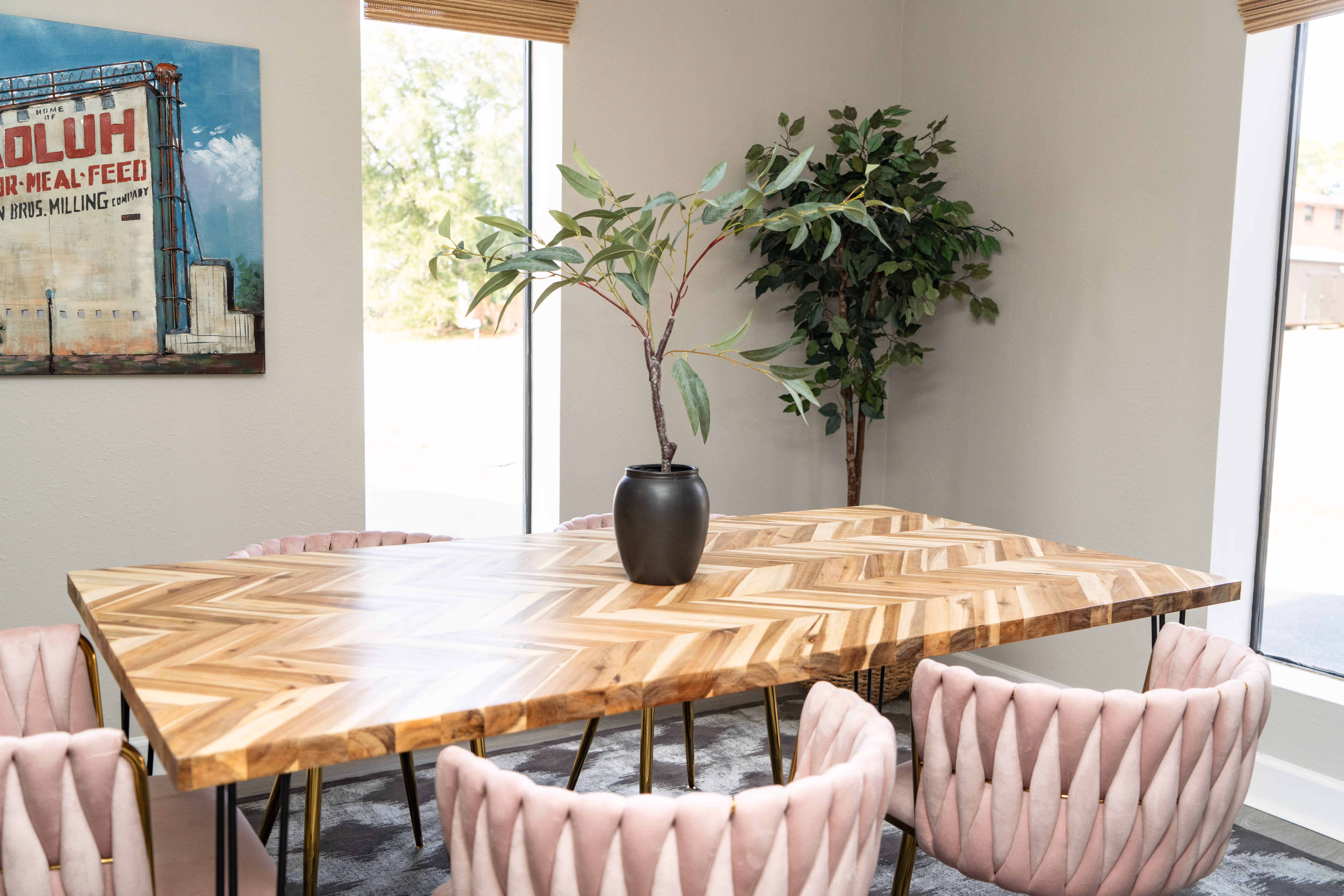 Modern dining table with a chevron-patterned wooden top surrounded by pink upholstered chairs and a black vase with green branches.