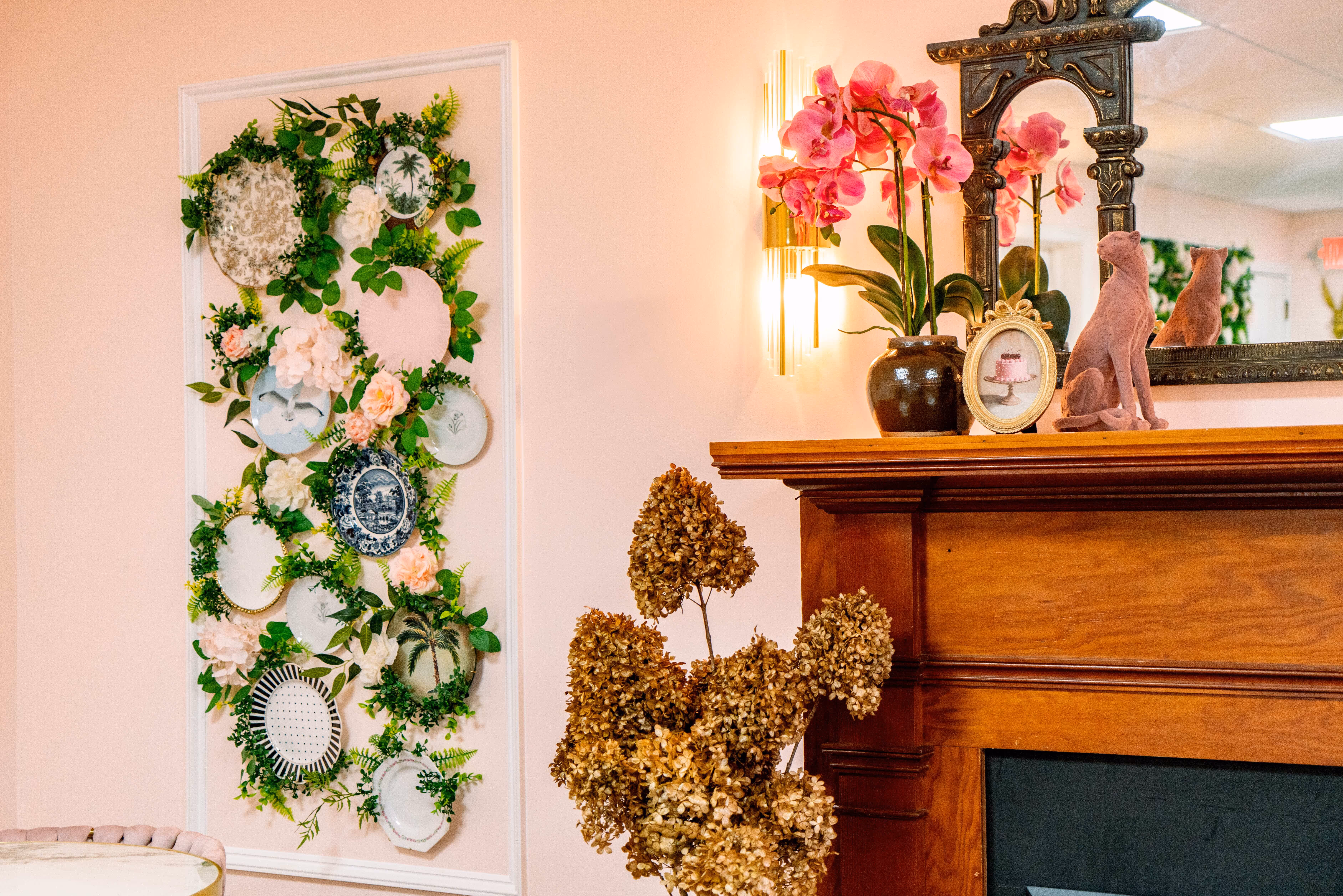 Wooden mantel with pink orchid in a brown vase, pink cat sculpture, framed cake photo, decorative wall plates with greenery, and dried hydrangea flowers.