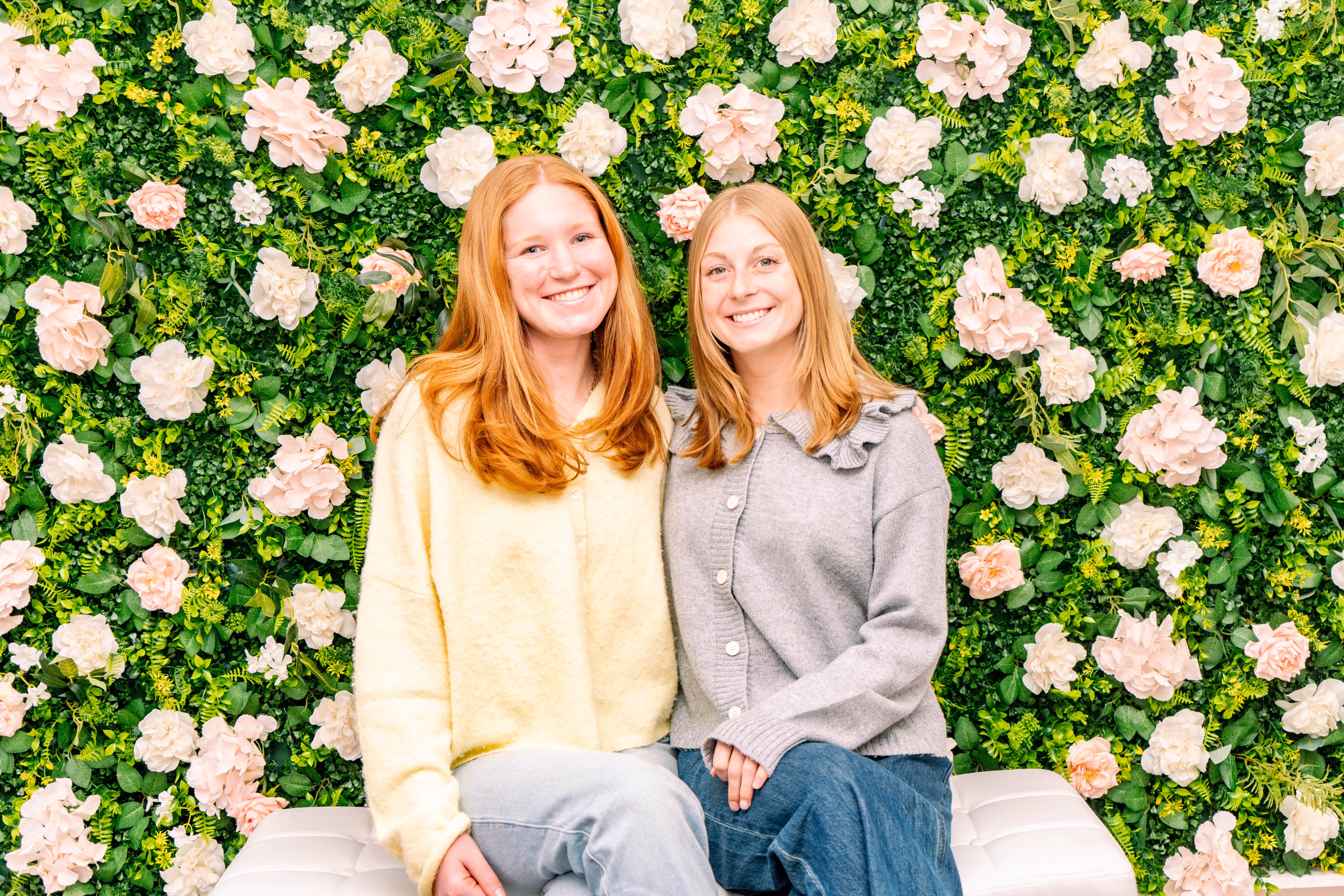 Two smiling women sitting side by side in front of a lush green wall decorated with pale pink and white flowers.