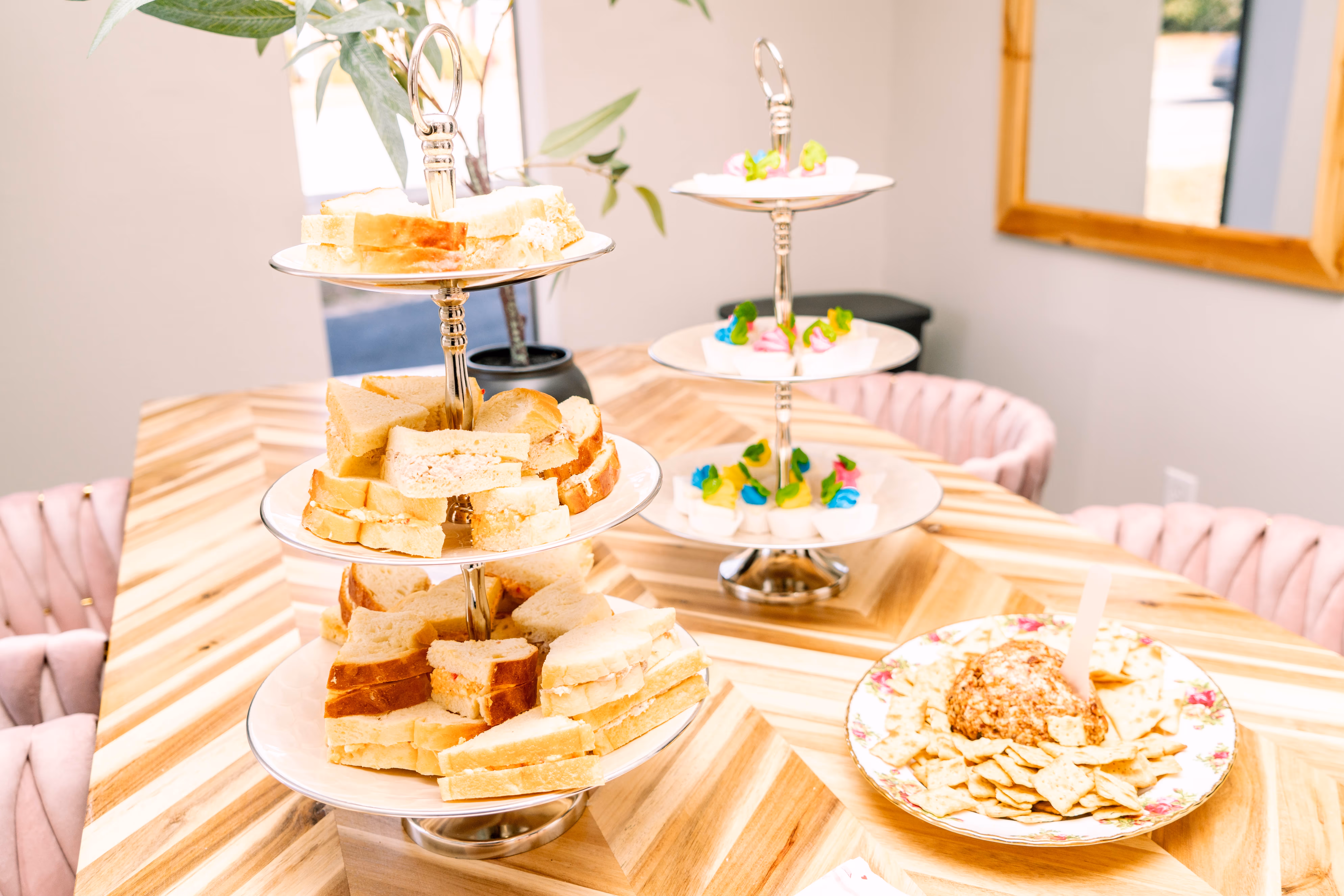 Three-tiered tray with assorted finger sandwiches and another three-tiered tray with colorful frosted cupcakes on a wooden table with a plate of cheese ball and crackers.