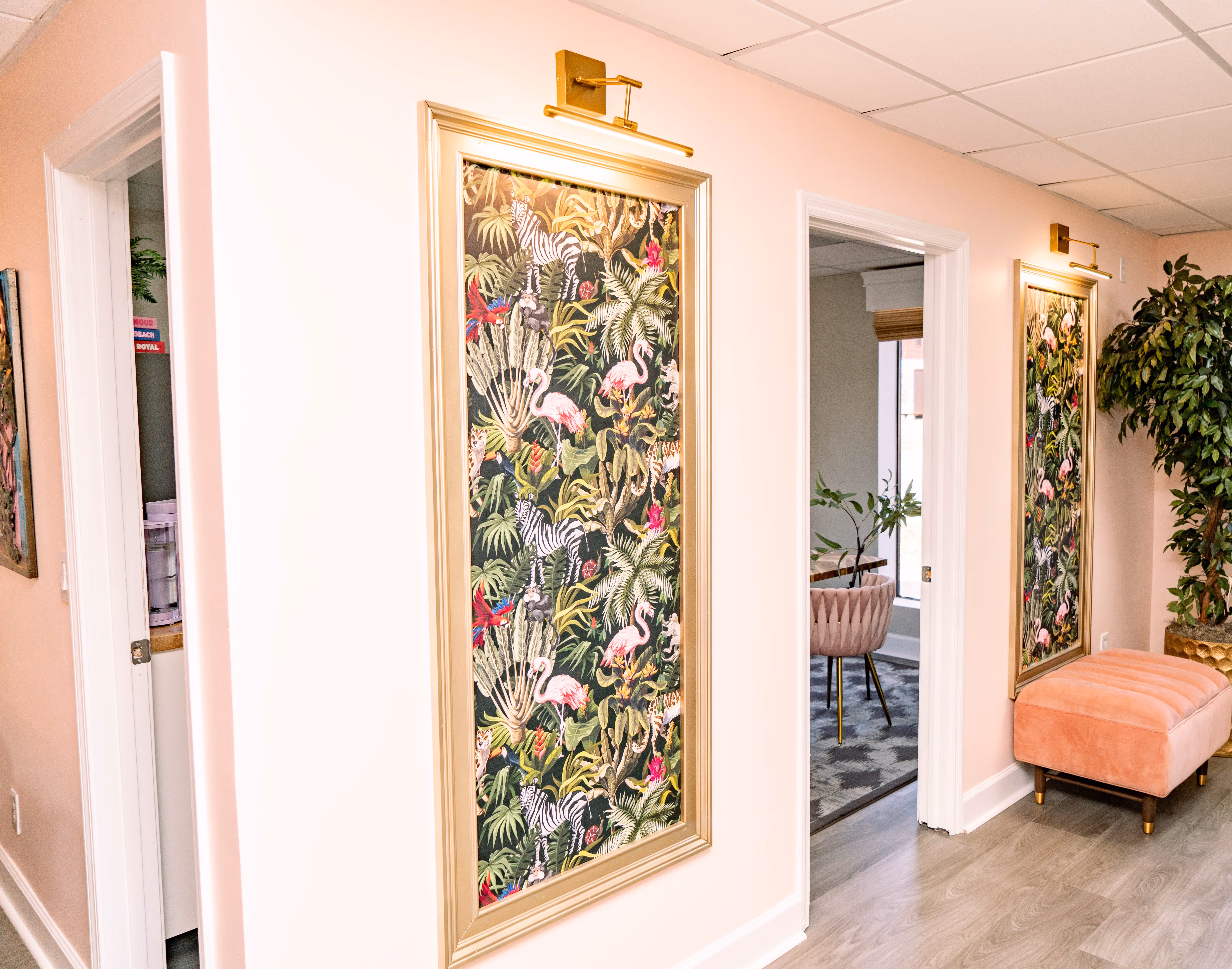 Interior hallway with light pink walls featuring two framed tropical prints with flamingos, a pink velvet bench, and a large potted plant.