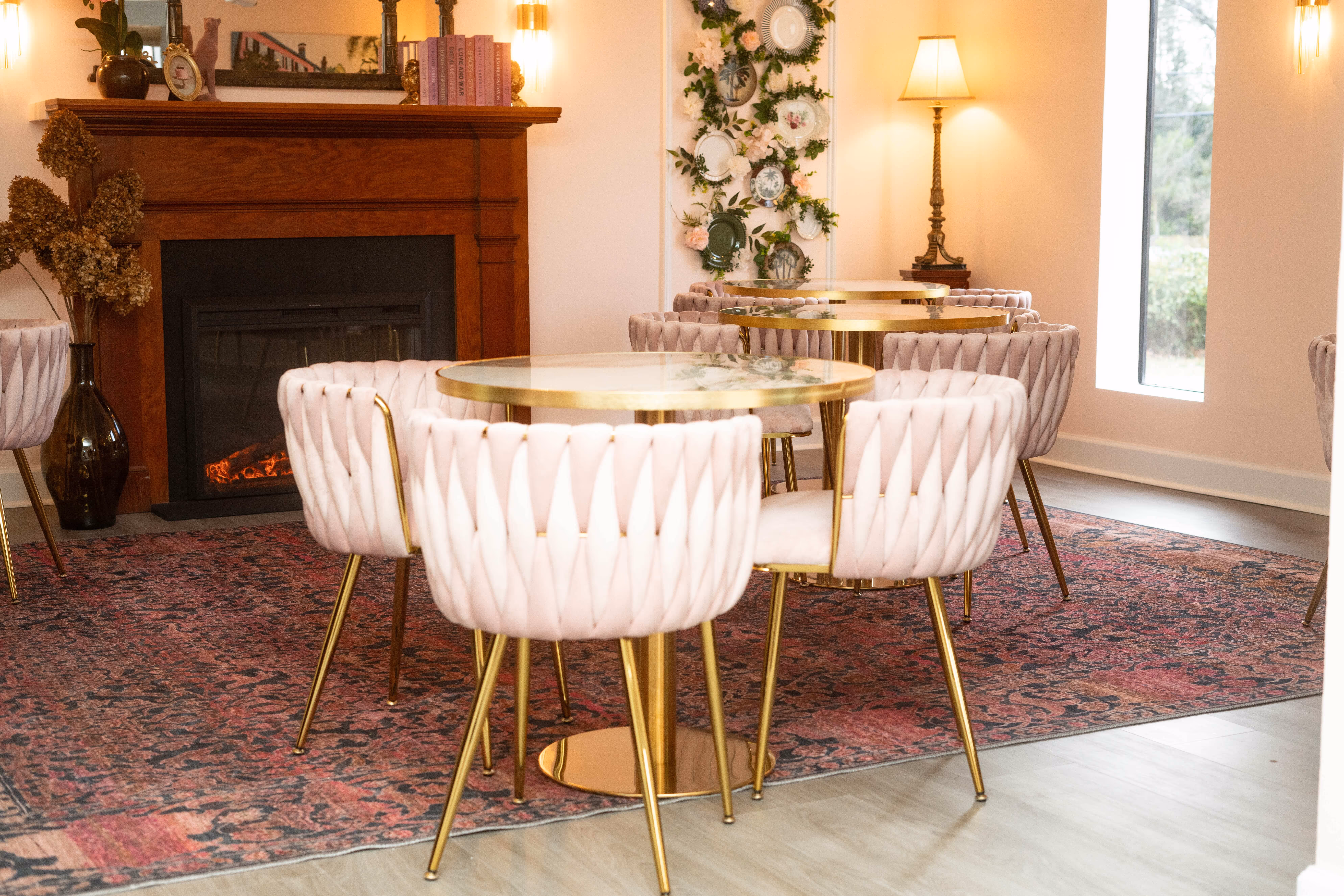 Cozy dining area with round gold tables and tufted pink chairs on a patterned rug in front of a wooden fireplace and floral plate wall decor.