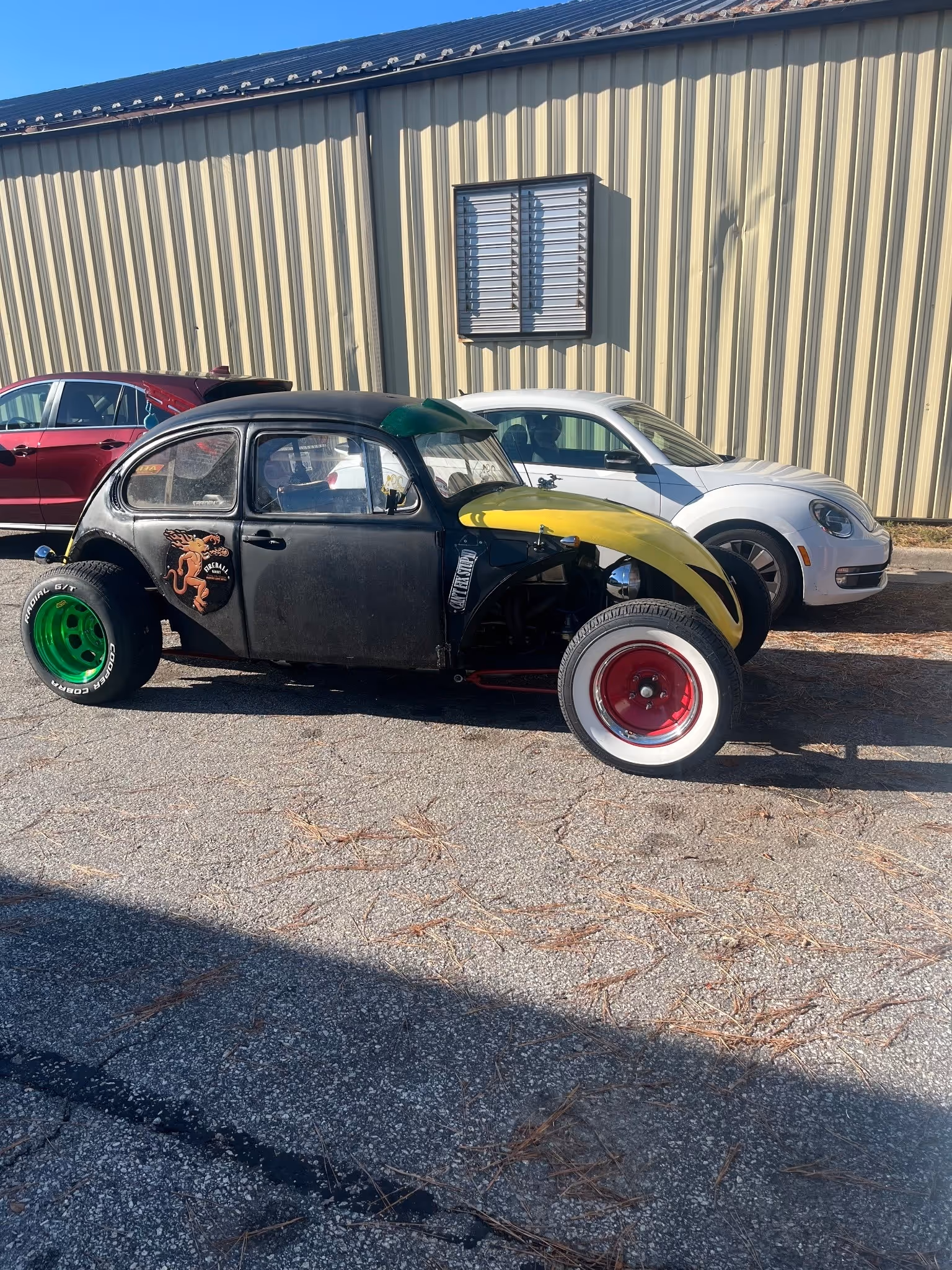 Black customized vintage Volkswagen Beetle with green rear wheels and whitewall front wheels parked next to a white and a red car beside a corrugated metal building.