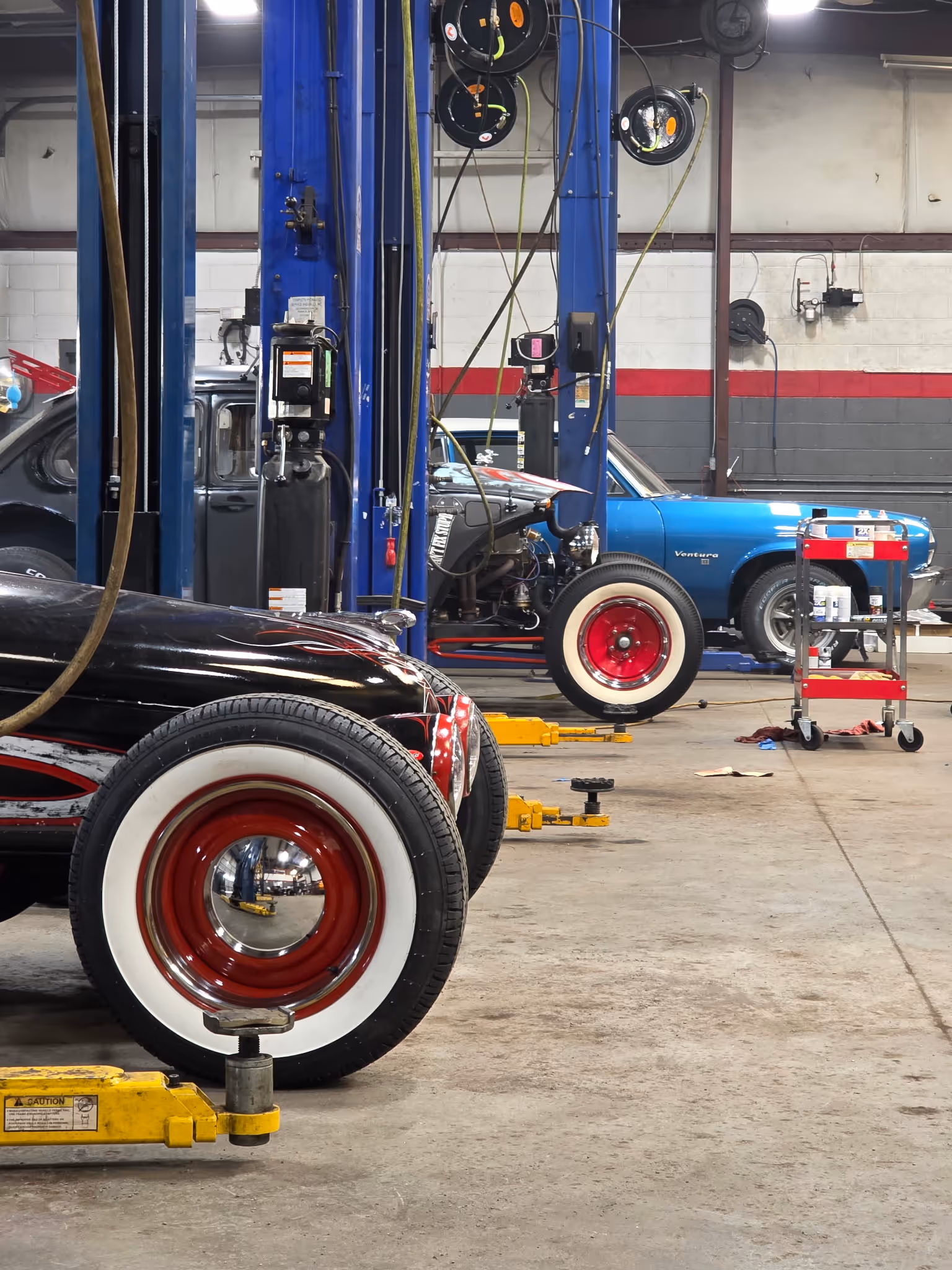 Classic cars with whitewall tires parked in an automotive repair shop with blue car lifts and tools.