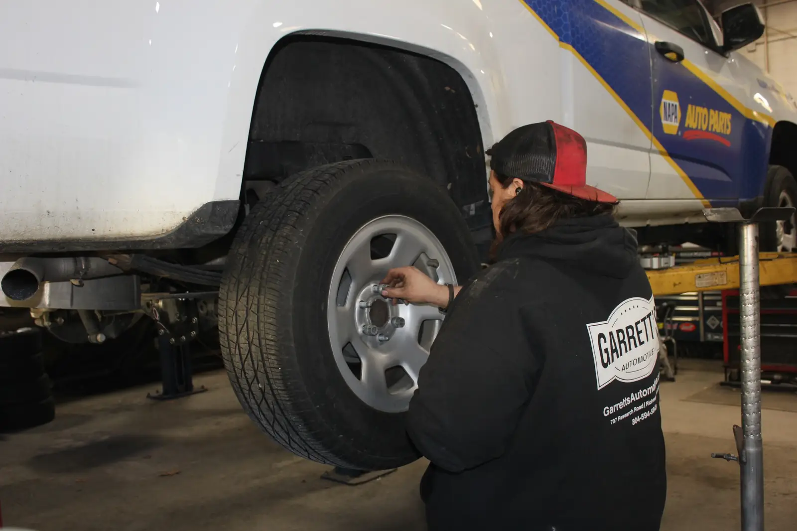 Mechanic wearing a black hoodie and red-black cap working on the wheel of a white vehicle with NAPA Auto Parts branding in a garage.