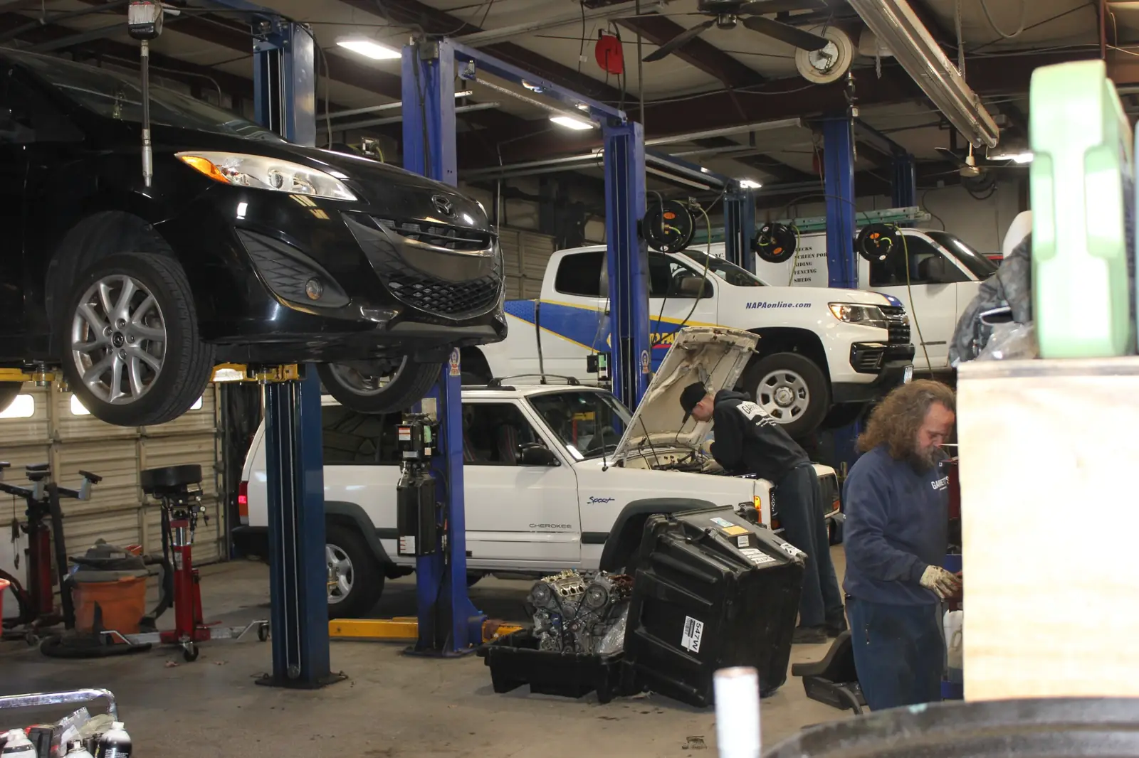 Auto repair shop with cars lifted on hoists, a mechanic inspecting a white Jeep Cherokee with its hood open, and another worker handling tools.