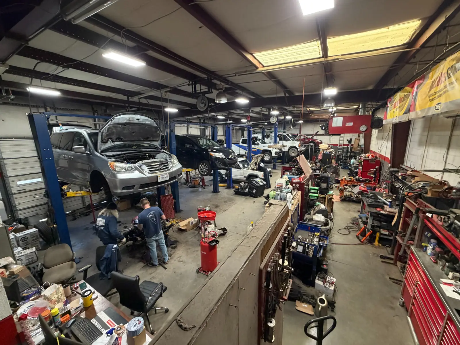 Interior of a busy auto repair shop with several cars on lifts, mechanics working, and tools and equipment scattered around.