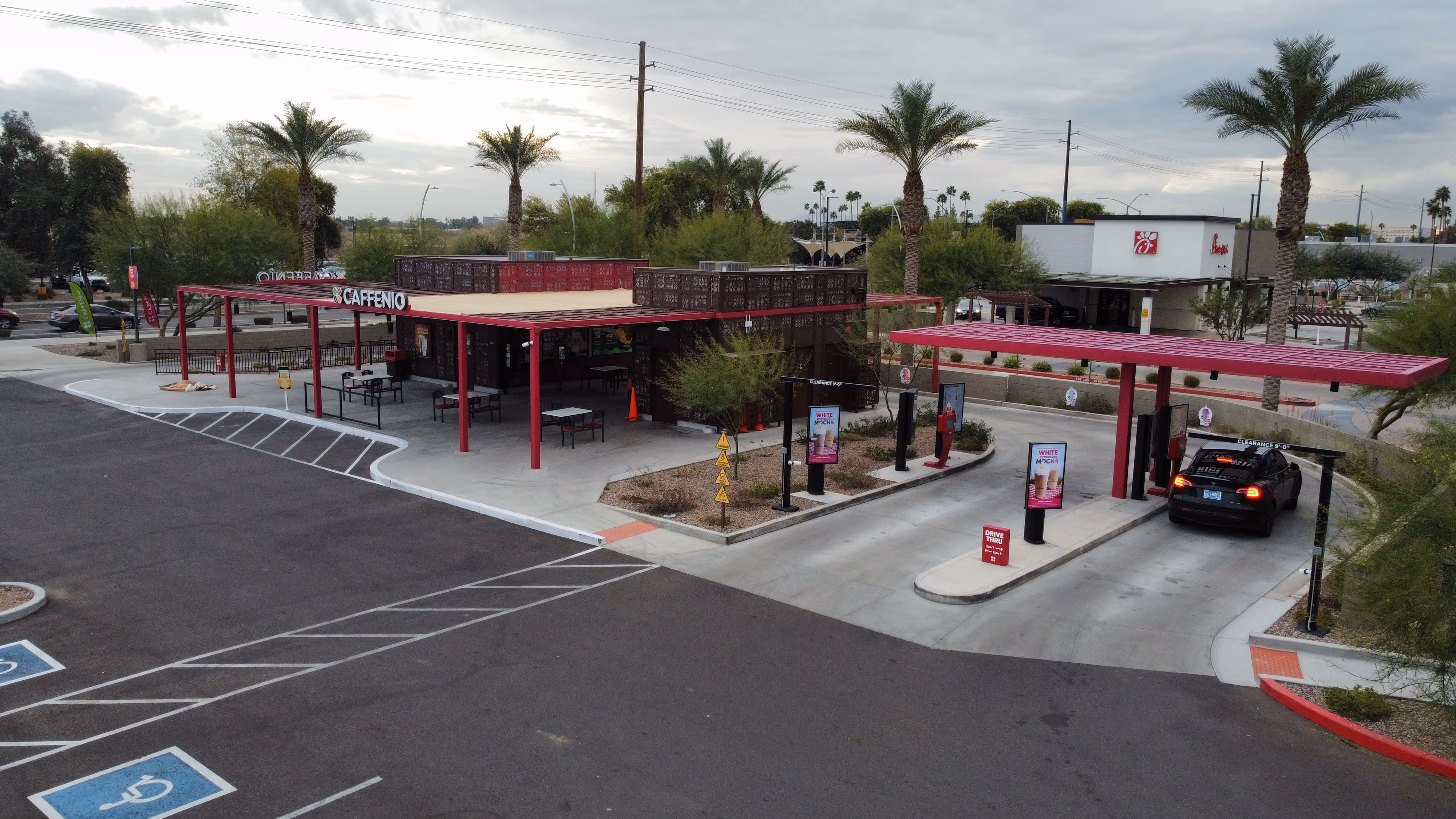 Aerial view of a Caffenio coffee shop with outdoor seating and a drive-thru lane next to a Chick-fil-A restaurant under cloudy sky.