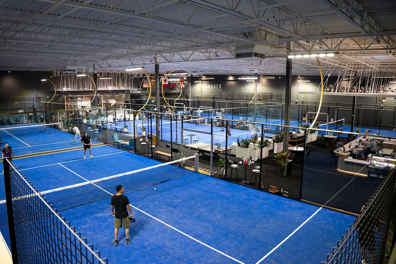 Indoor padel club with multiple blue courts, players actively playing, and a seating area beside the courts.