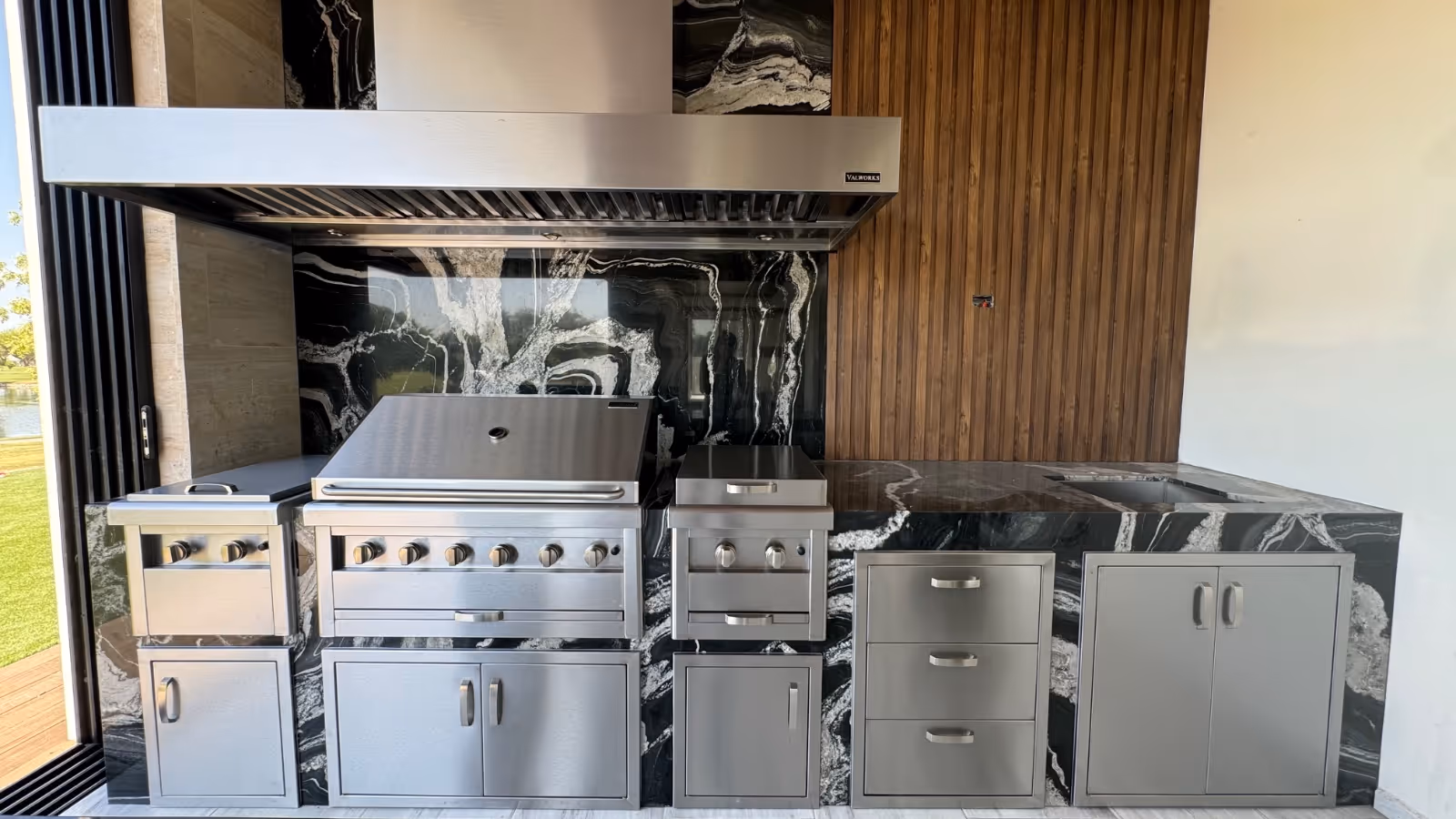 Modern outdoor kitchen setup featuring stainless steel grill, stove, storage cabinets, and black marble countertop with wooden wall background.