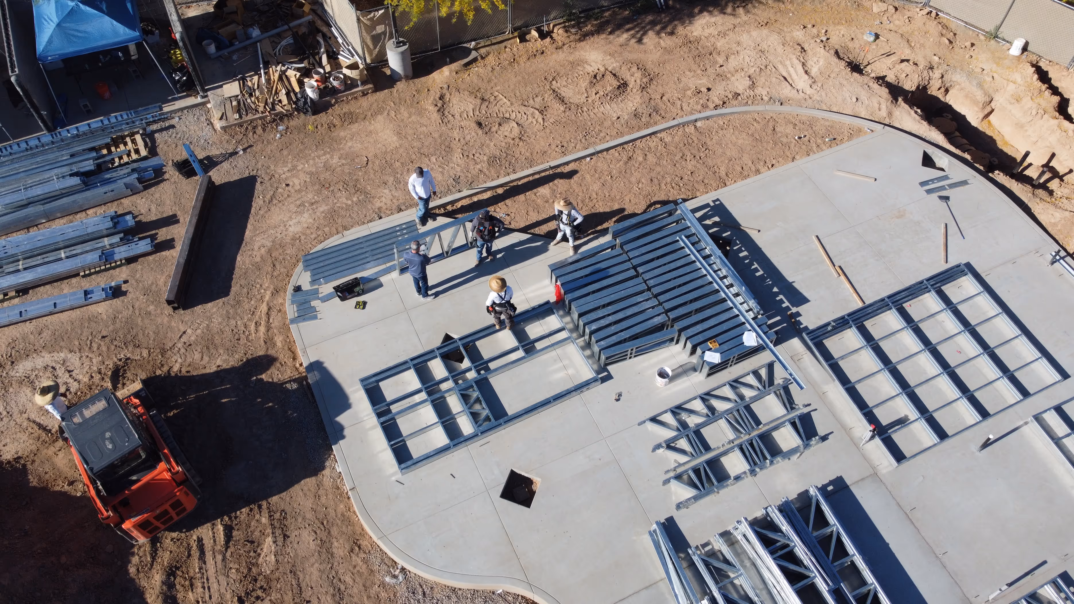 Aerial view of construction workers assembling steel frames on a concrete foundation at a construction site with equipment and dirt surrounding.