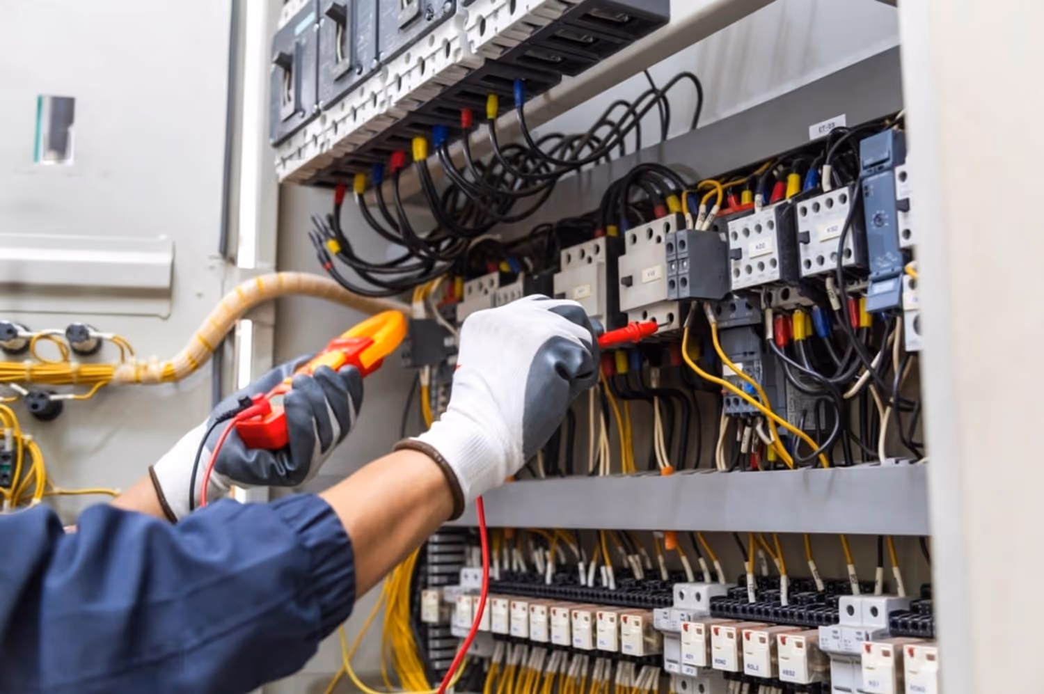 Electrician wearing gloves testing wires inside an electrical panel with a multimeter.