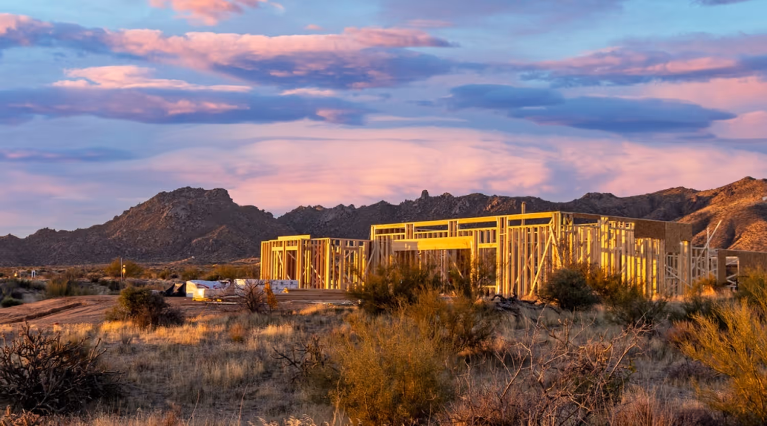 Wooden frame structure of a house under construction in a dry, shrub-filled landscape with mountains and a colorful sunset sky.
