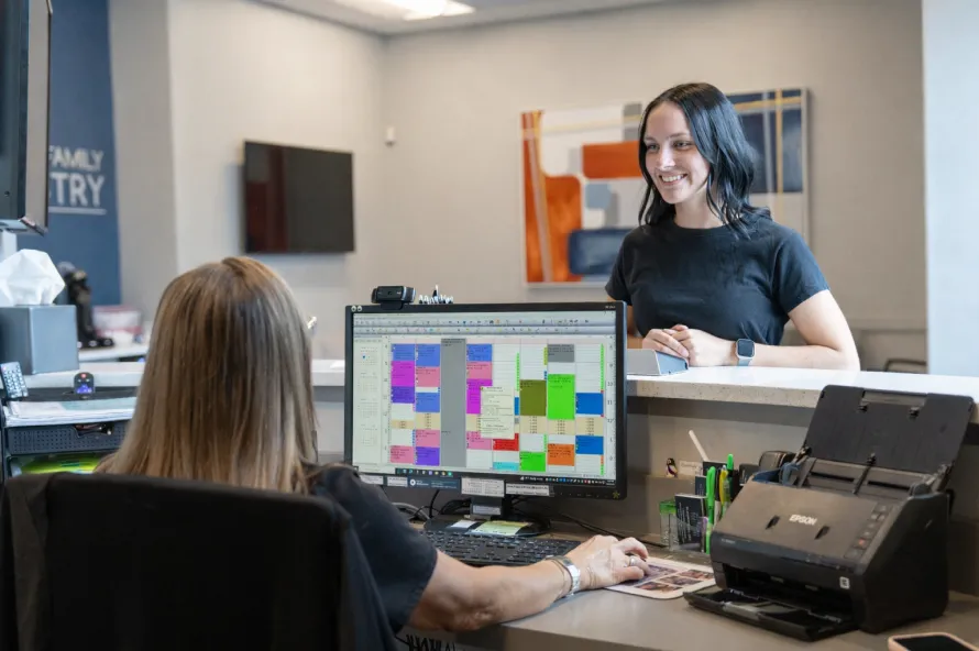 Receptionist sitting at a desk with a colorful calendar on the computer screen, smiling and assisting a woman standing at the counter.