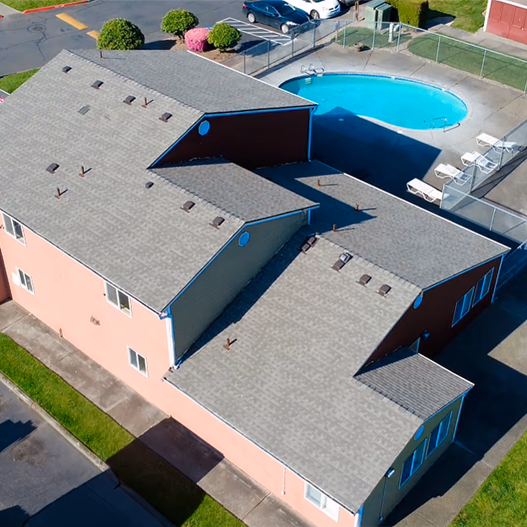 Aerial view of a townhouse complex with grey roofs, a fenced oval swimming pool, poolside lounge chairs, and parked cars nearby.