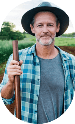 Middle-aged man in a blue plaid shirt and hat holding a wooden stick in a green outdoor setting.