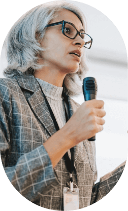 Middle-aged woman with gray hair and glasses speaking into a microphone while wearing a plaid blazer and ID badge.