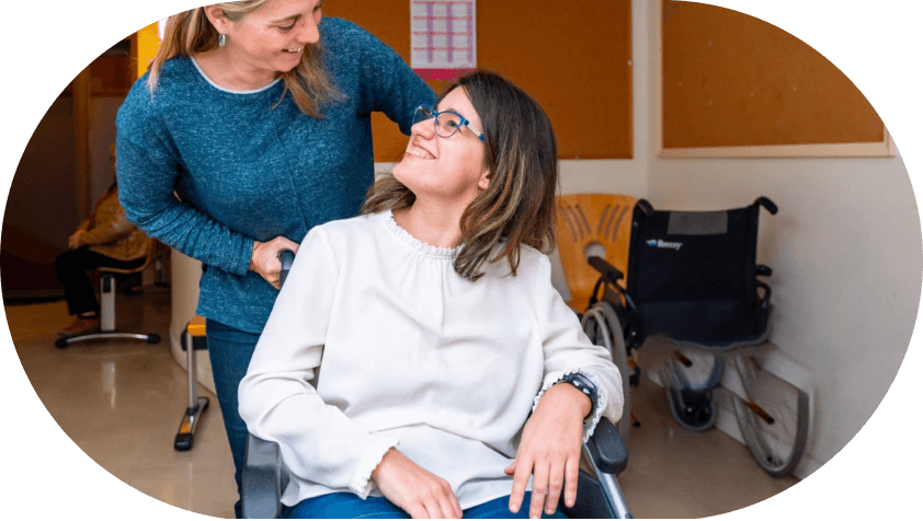 A smiling woman in a wheelchair looks up at another woman standing behind her, indoors near another empty wheelchair.