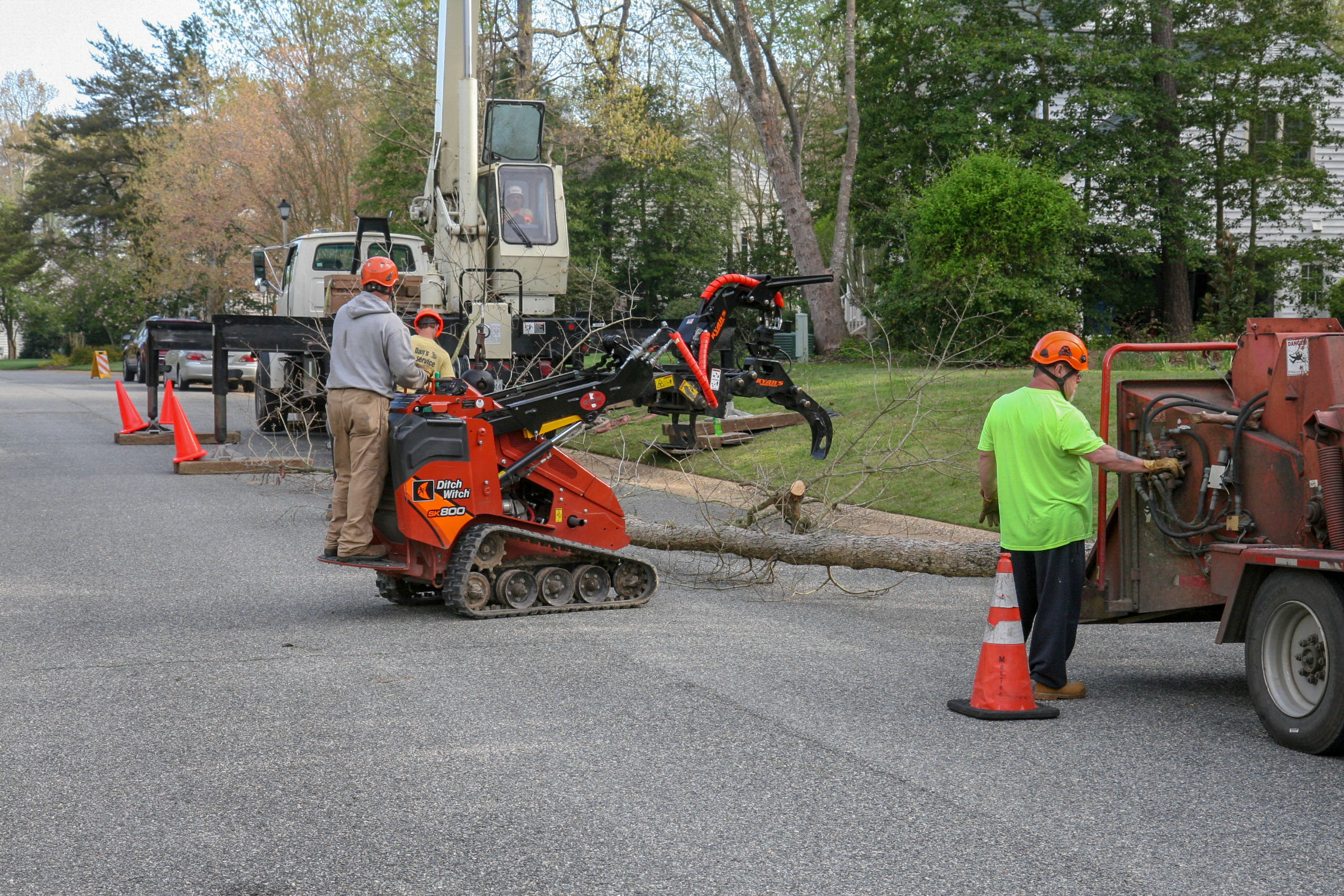 Workers with safety helmets operate machinery to remove and chip a tree trunk on a residential street.