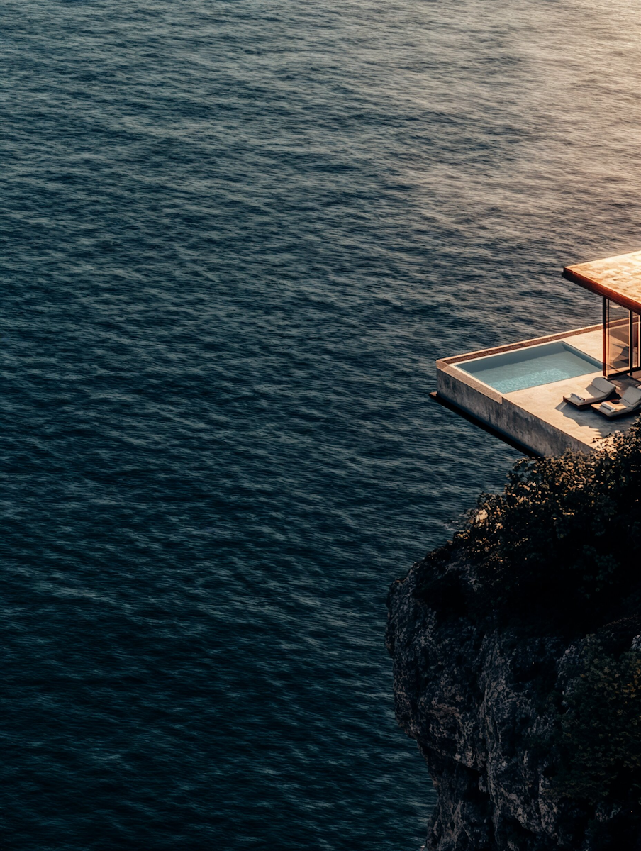 Modern villa with a small pool and lounge chairs perched on a rocky cliff overlooking the ocean during sunset.