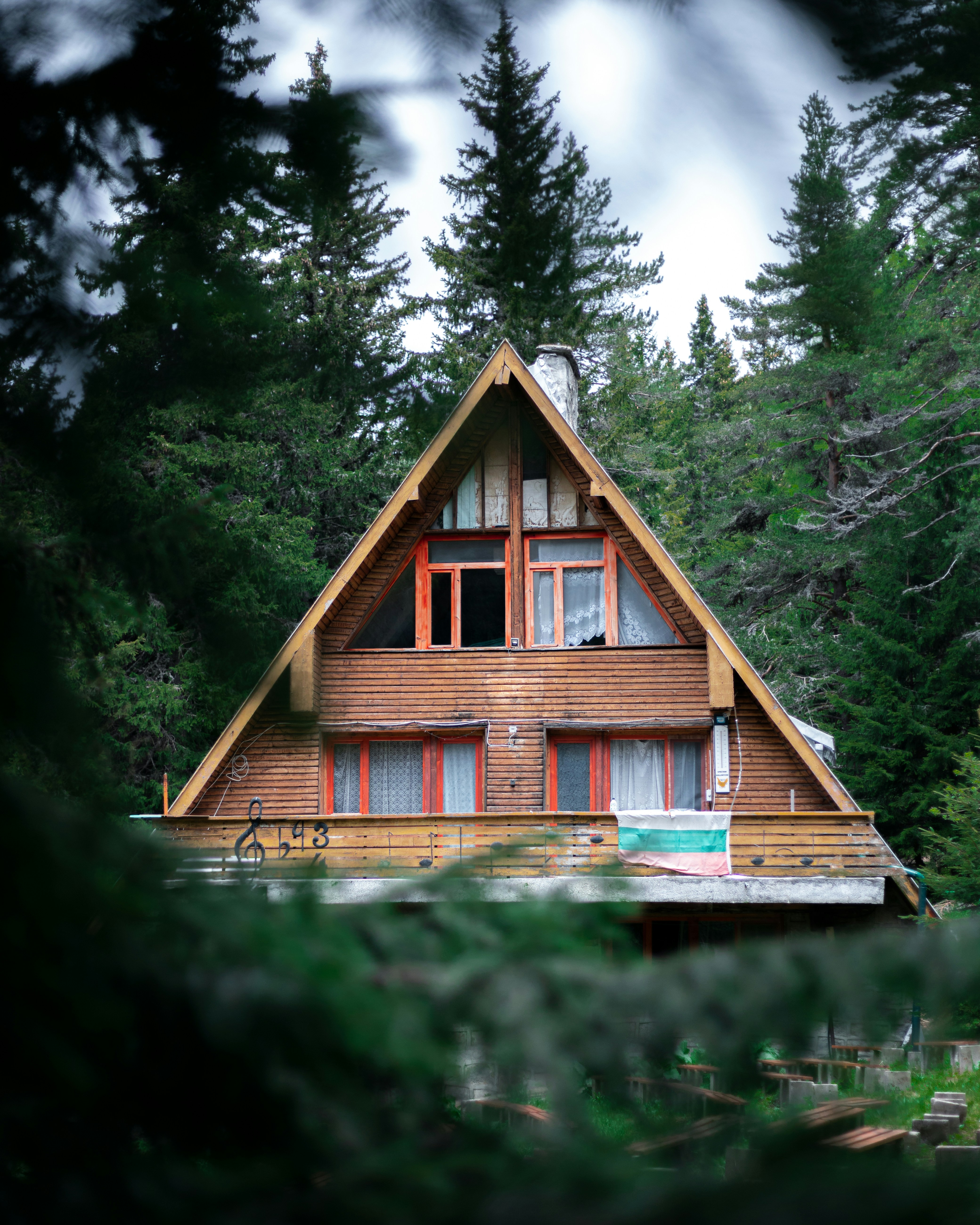 Wooden A-frame house with red-trimmed windows surrounded by dense evergreen forest.