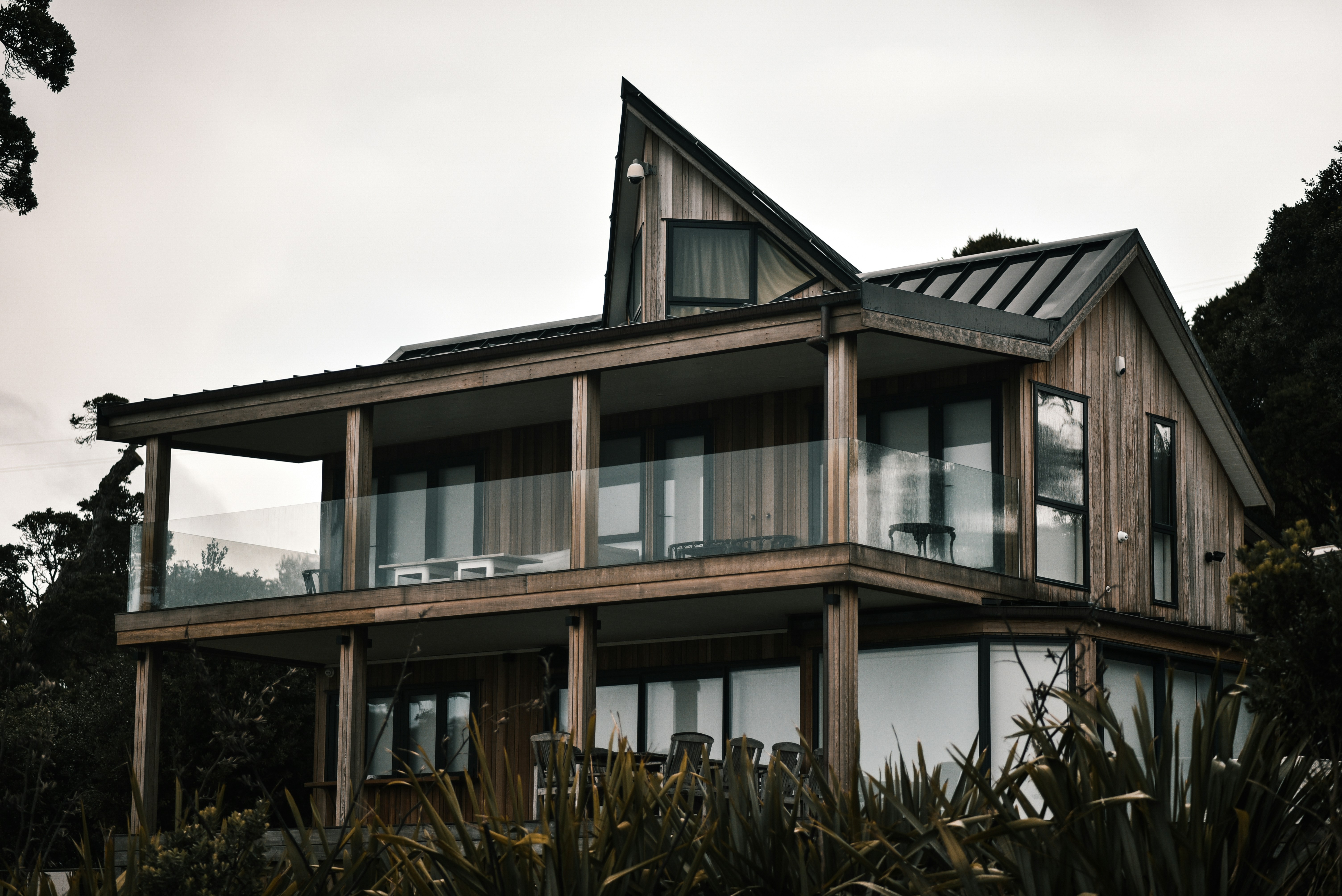 Modern two-story wooden house with large glass balcony and triangular roof surrounded by trees and plants.