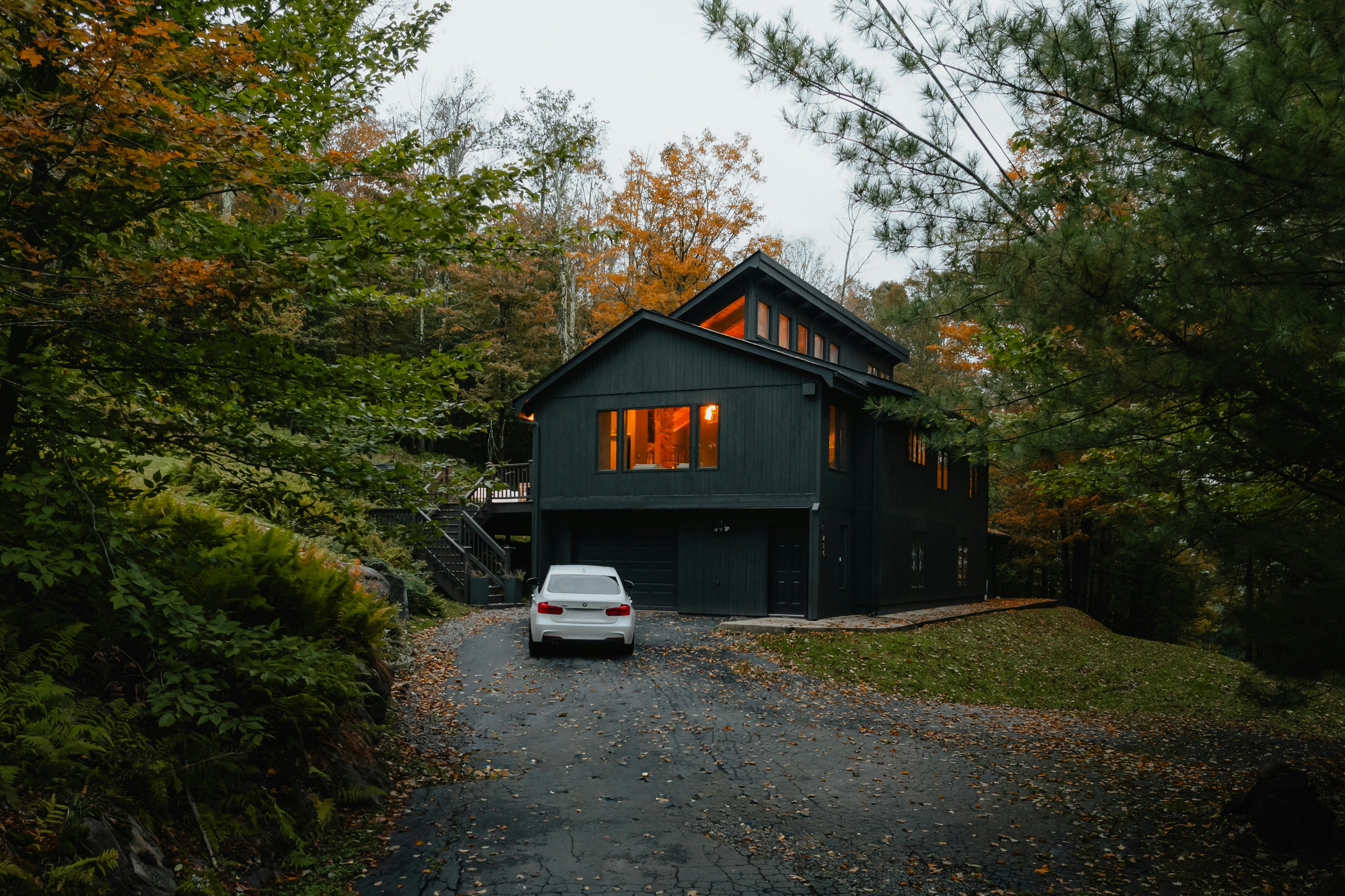 Modern dark house with illuminated windows surrounded by autumn trees and a white car parked on the driveway.