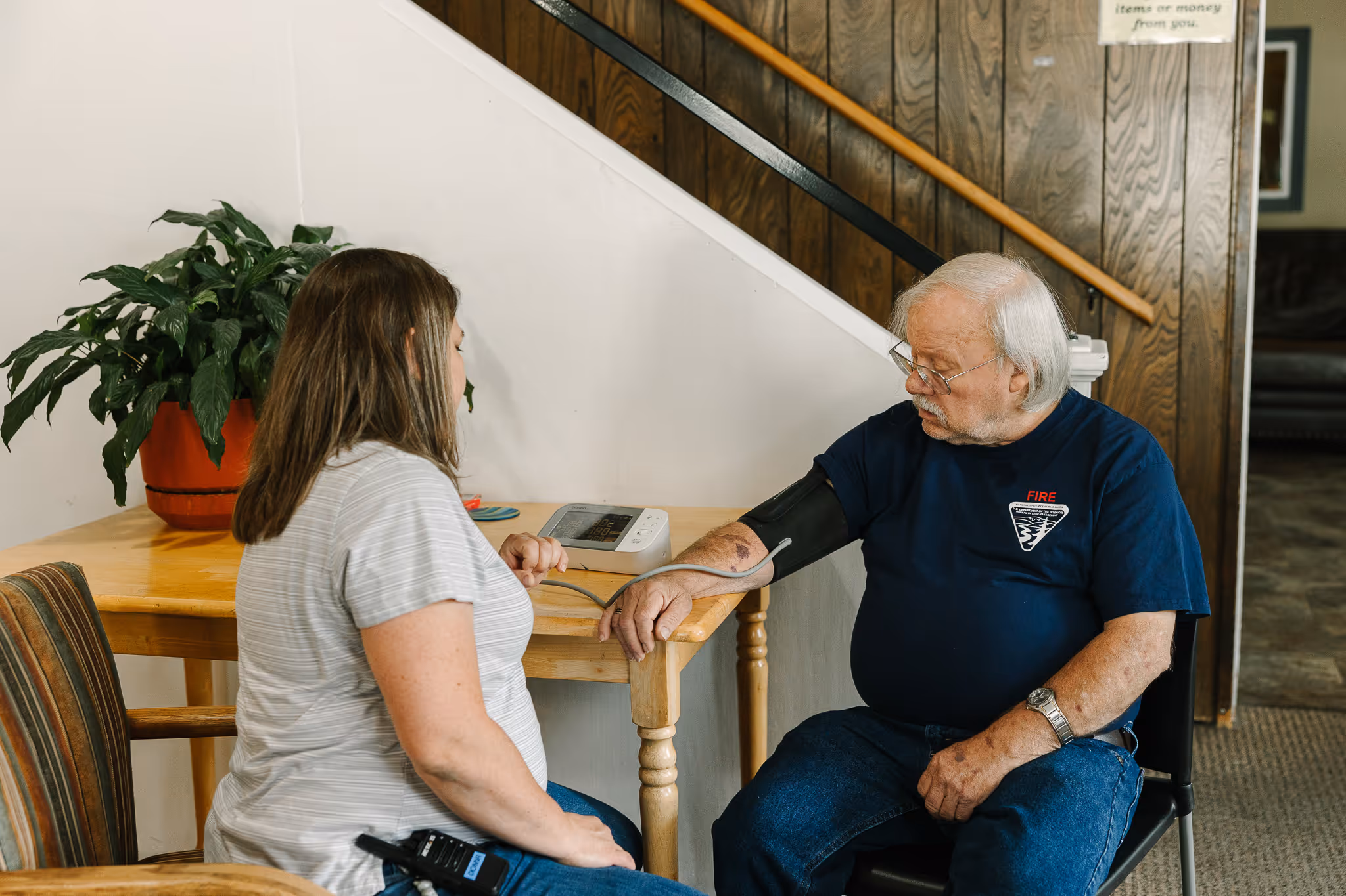 Woman measuring elderly man's blood pressure with digital monitor at a wooden table indoors.
