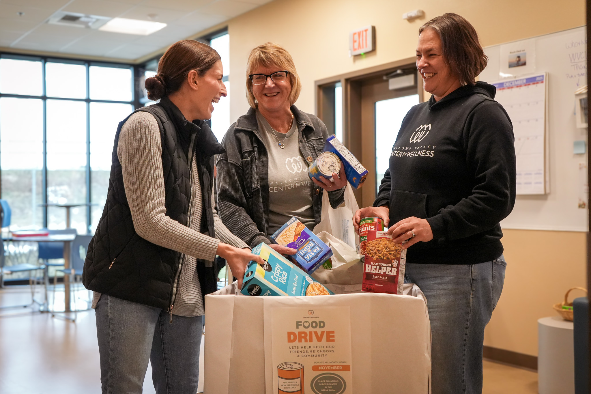 Three women smiling while placing food items into a large box labeled Food Drive at a wellness center.