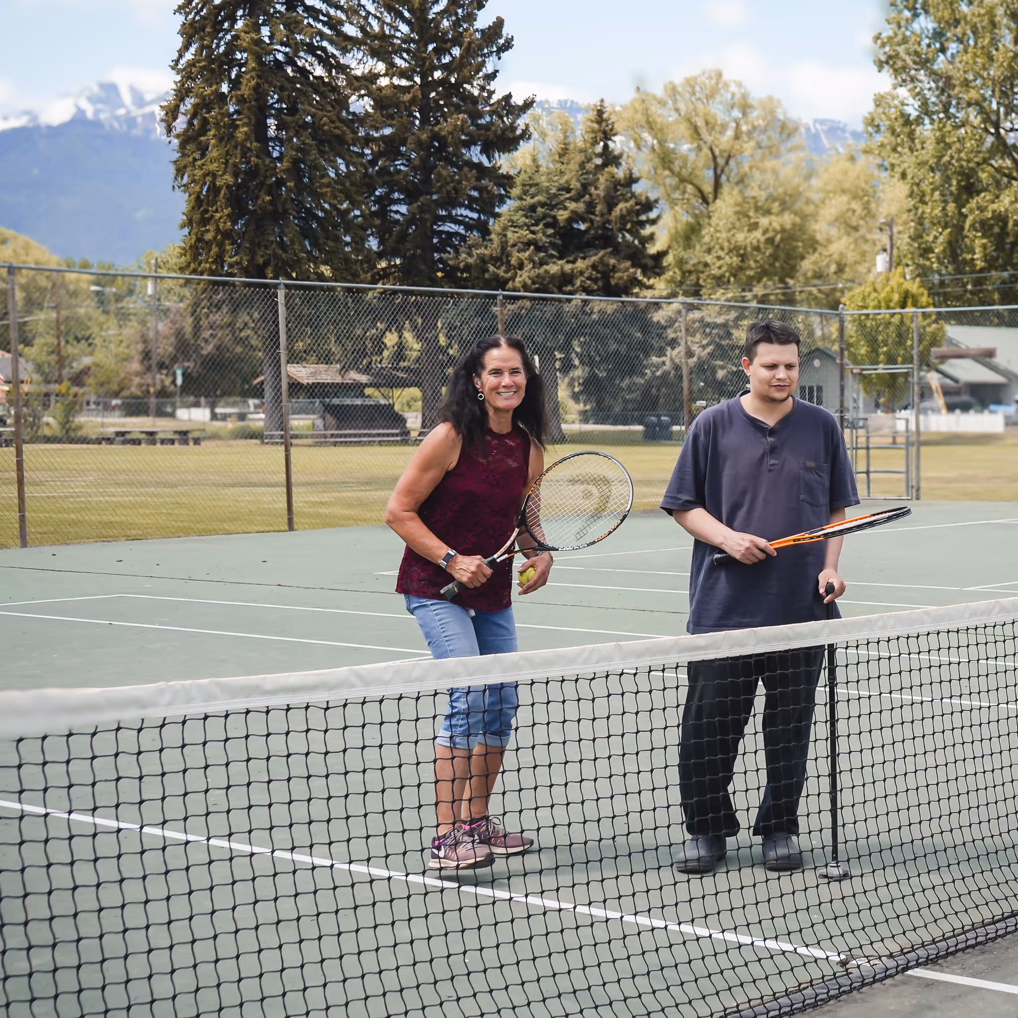 Woman and man holding tennis rackets standing on an outdoor tennis court near the net.