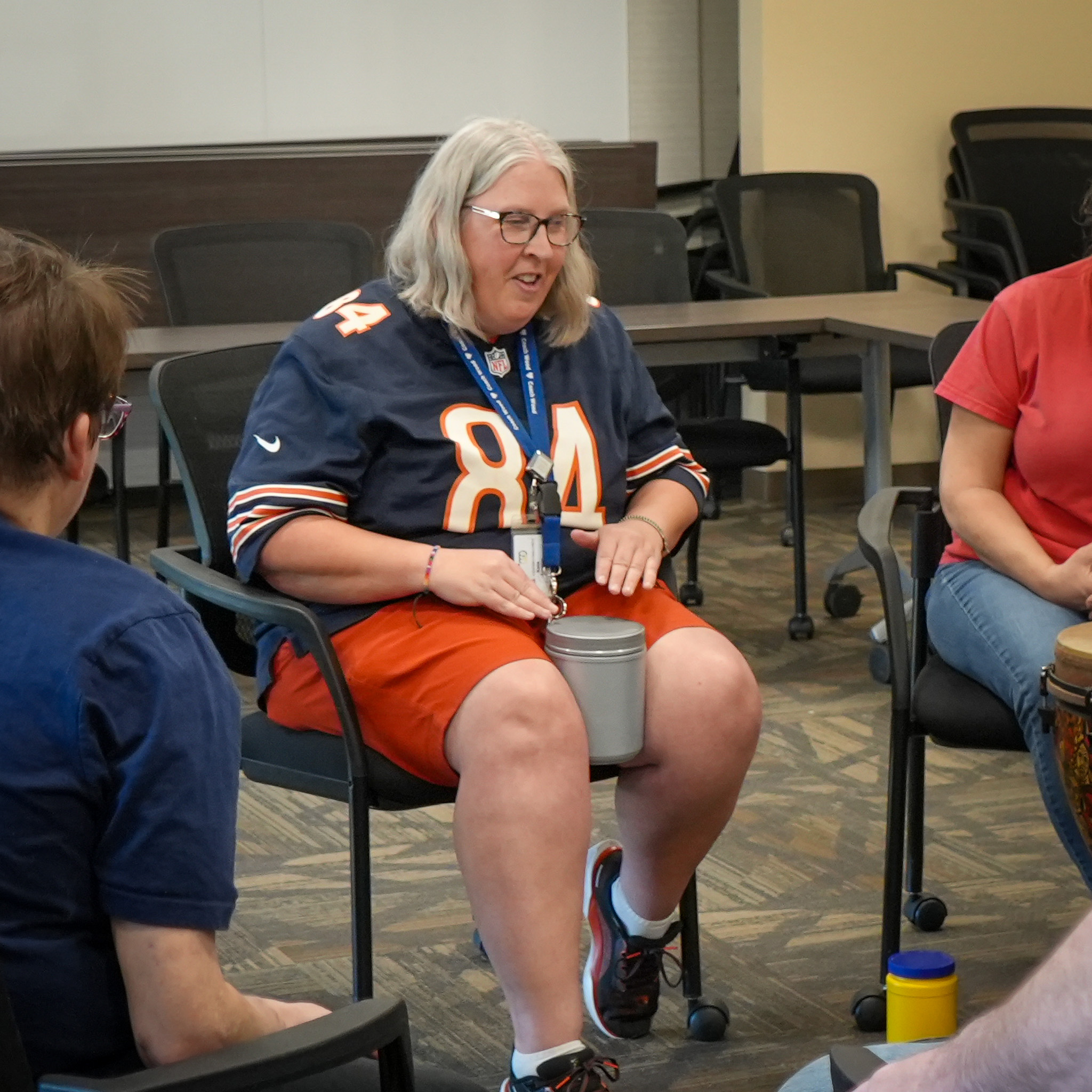 Woman sitting in a meeting room playing a small hand drum on her lap with other participants.