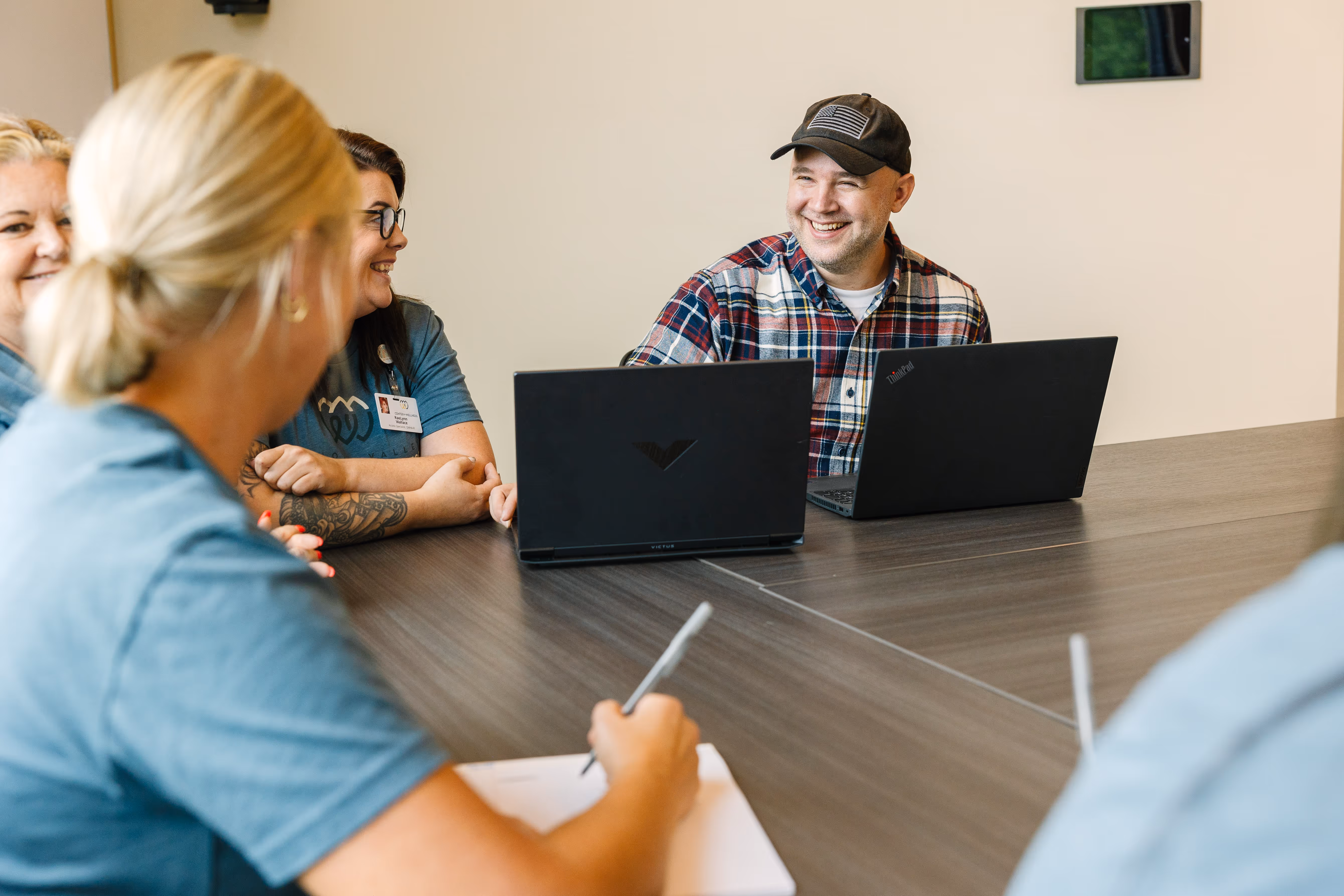 Smiling man in a cap and plaid shirt at a table with three women during a meeting with laptops.