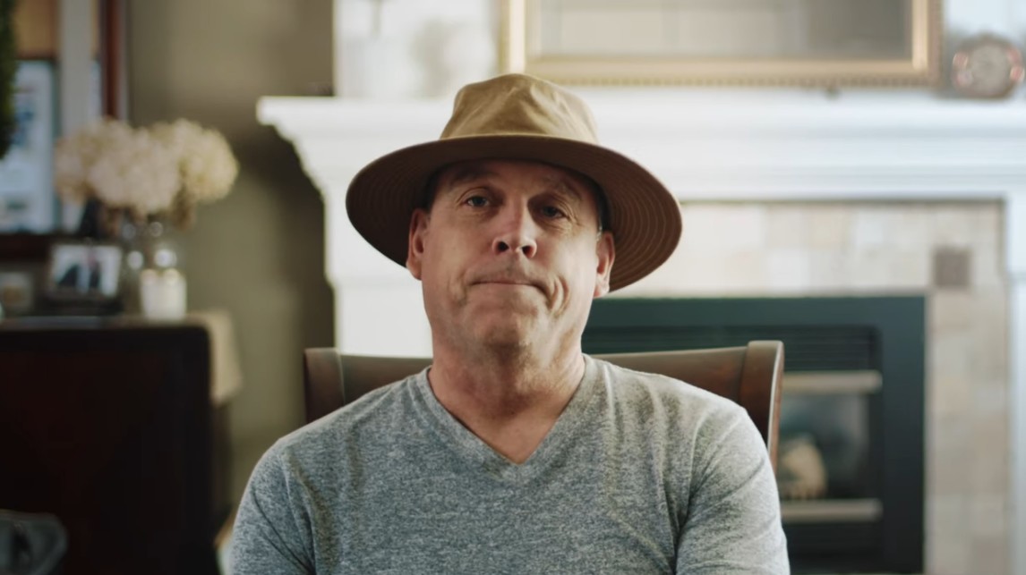 Man wearing a brown hat and gray shirt sitting indoors with a fireplace in the background.