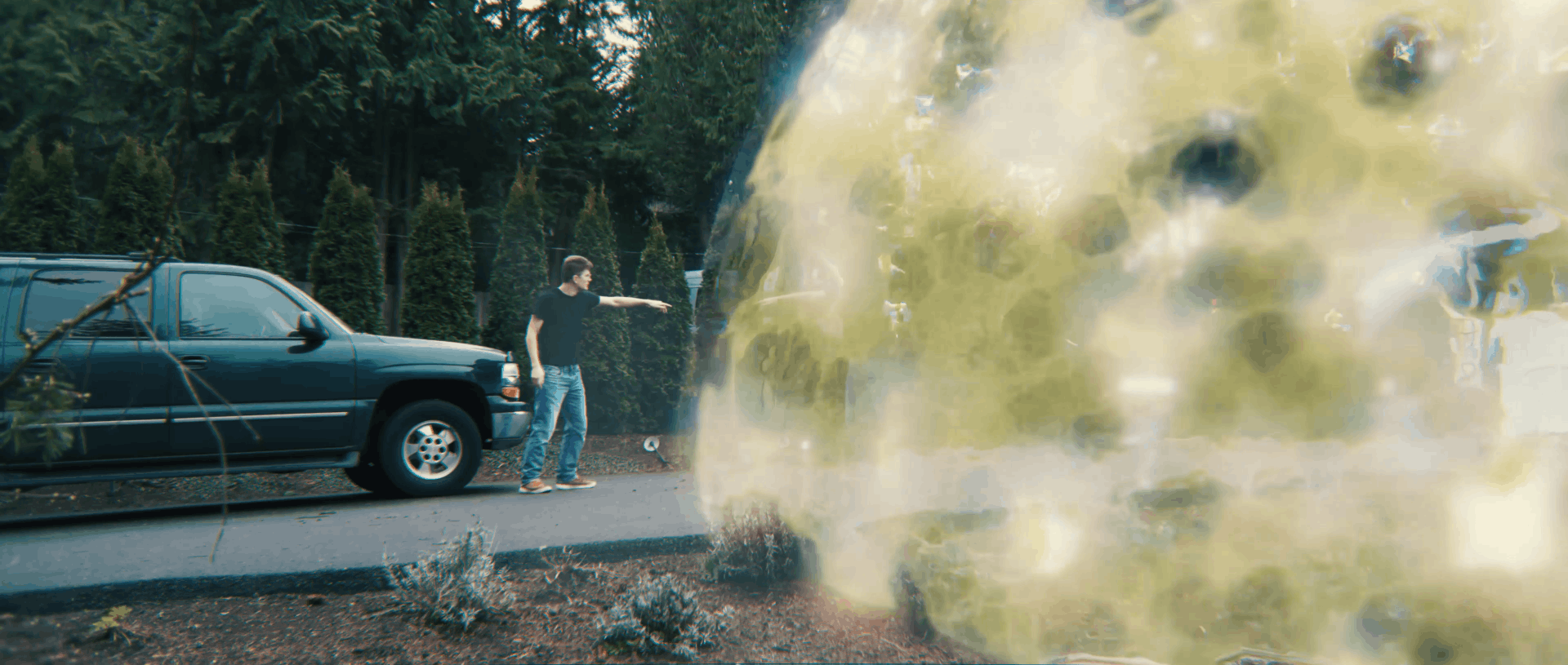 A man in jeans and a black shirt stands on a driveway pointing toward a large glowing, translucent yellow-green energy sphere next to a parked black SUV.
