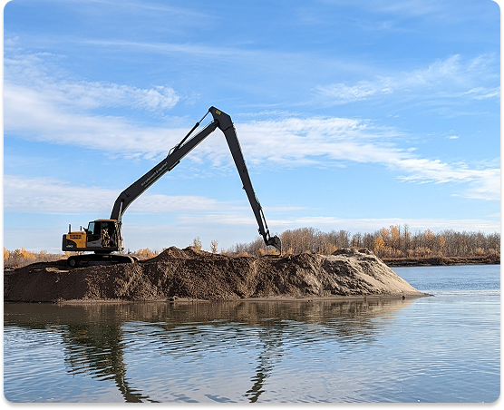 Excavator on a mound of dirt next to a body of water under a blue sky with scattered clouds.