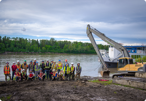 Group of construction workers wearing safety gear posing by a riverbank next to an excavator under a partly cloudy sky.