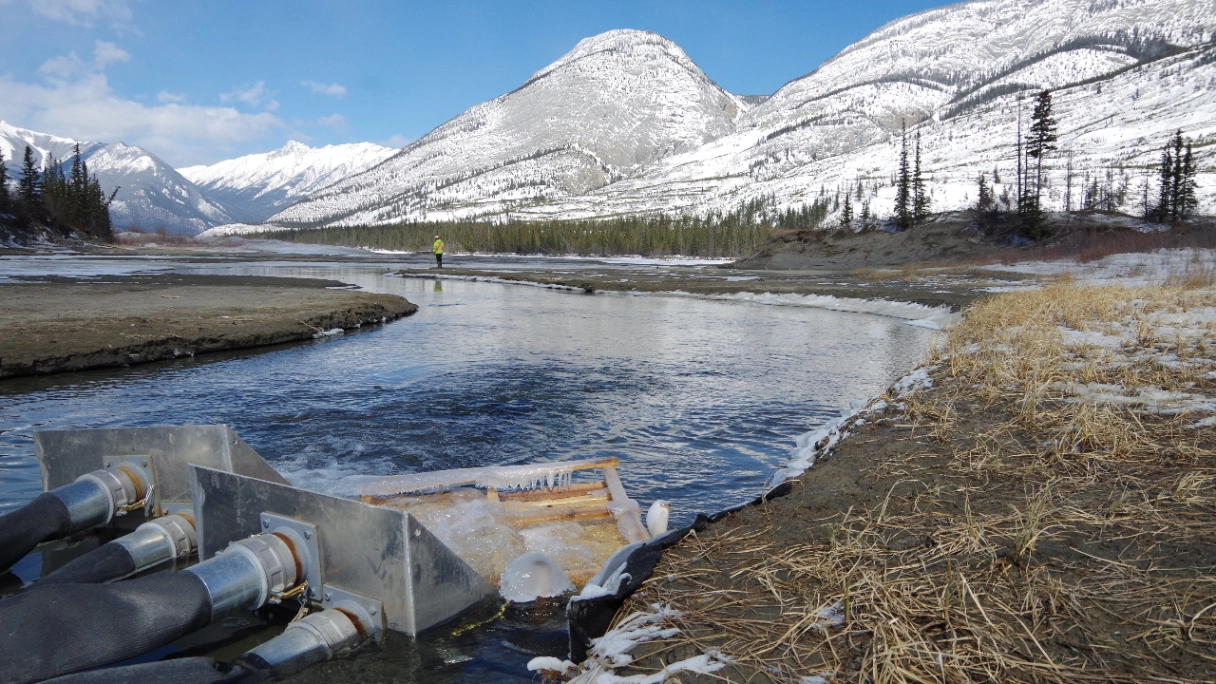 Snow-covered mountains and a river with pipes flowing water into it under a clear blue sky.